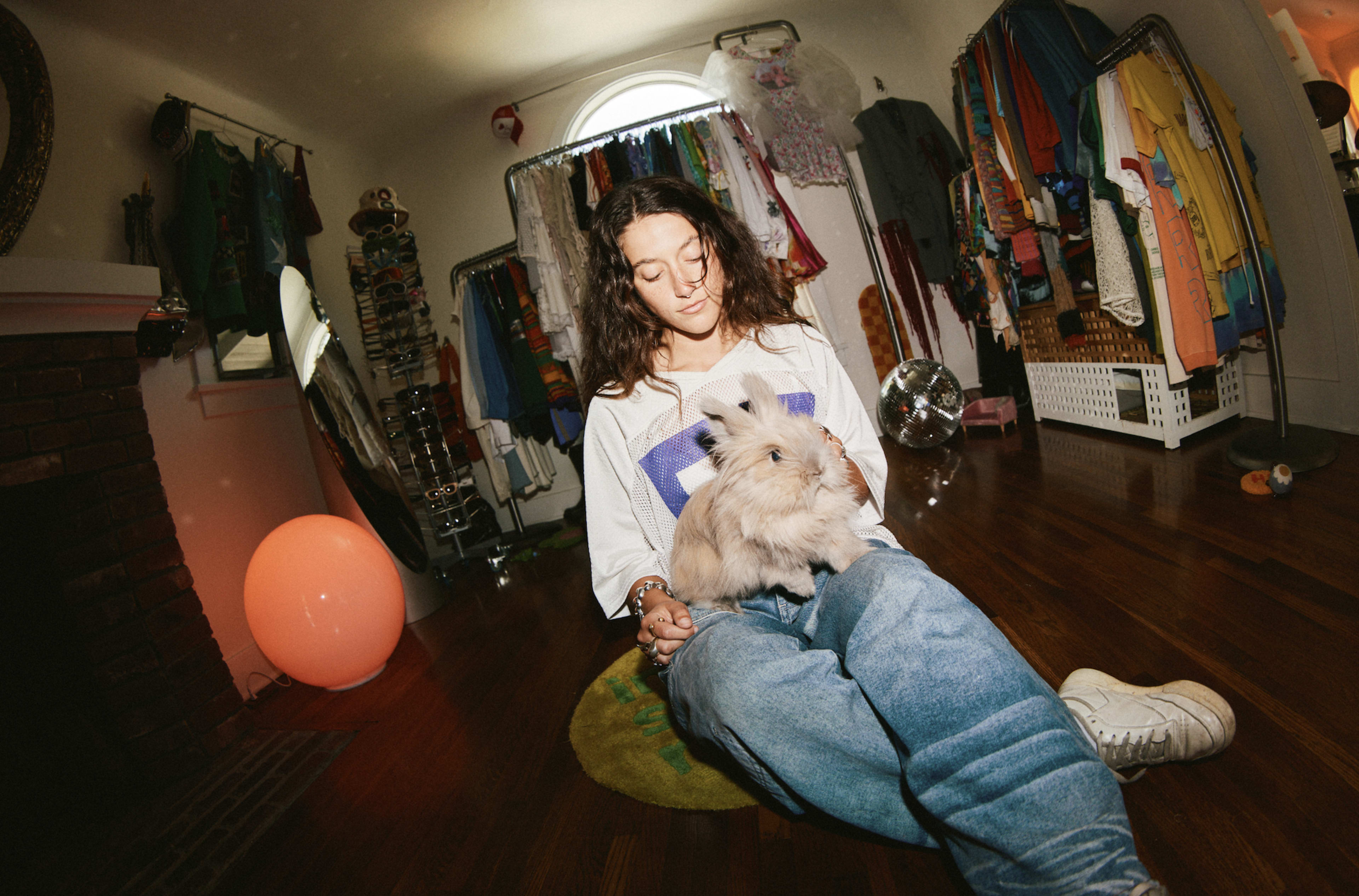 A person wearing casual clothing sits on the floor holding a rabbit, surrounded by clothing racks and a softly lit room.