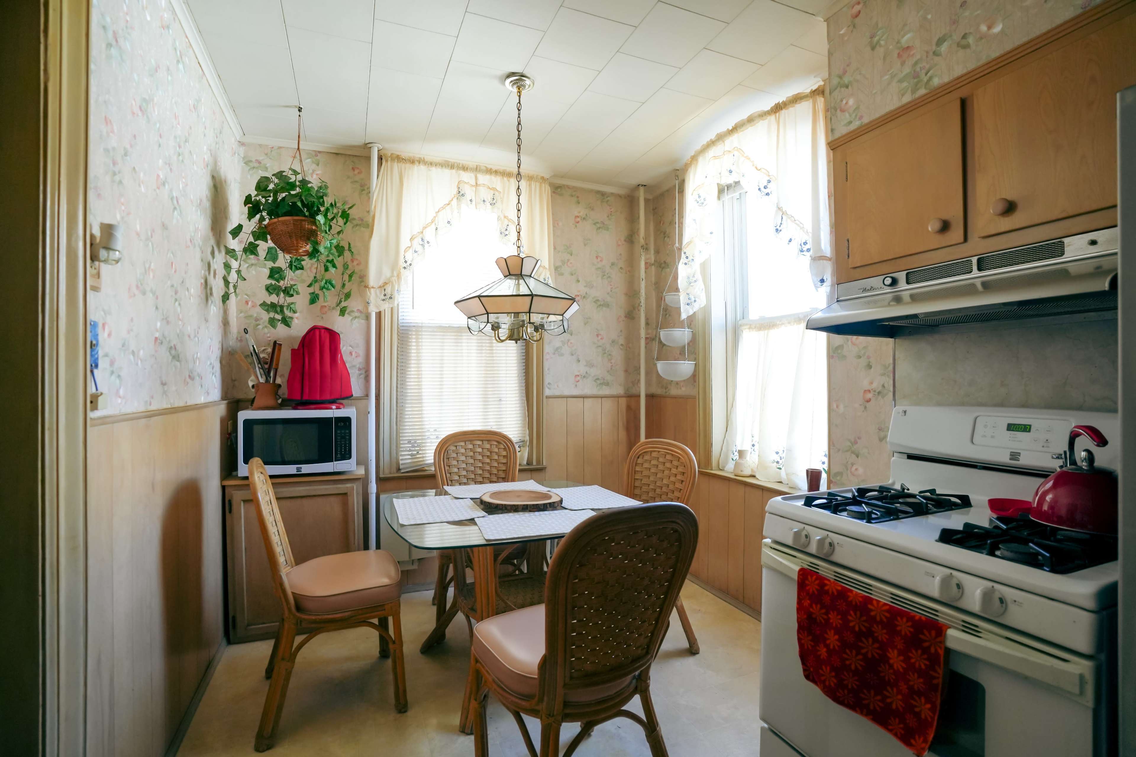 A small kitchen features a table with four chairs, a gas stove, a microwave, and floral wallpaper, illuminated by natural light from two windows.