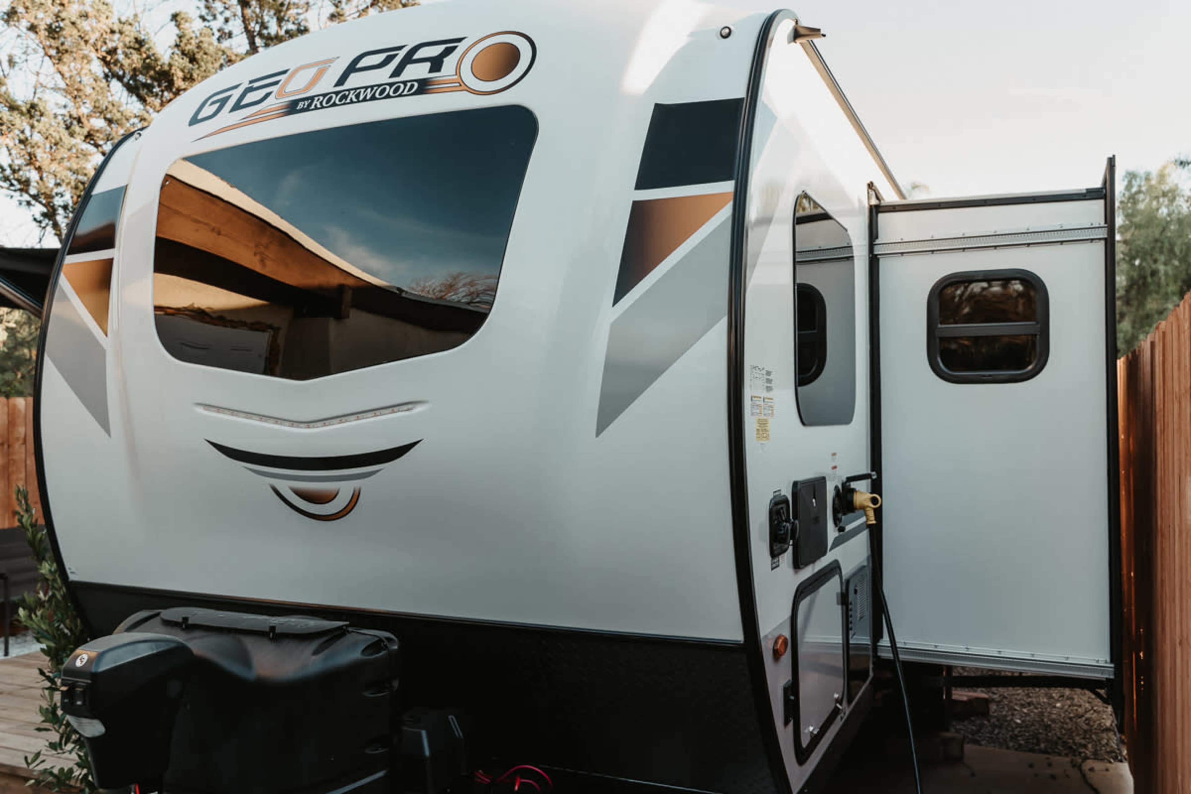 A Rockwood Geo Pro travel trailer parked beside a wooden fence, with the entrance door and window visible.