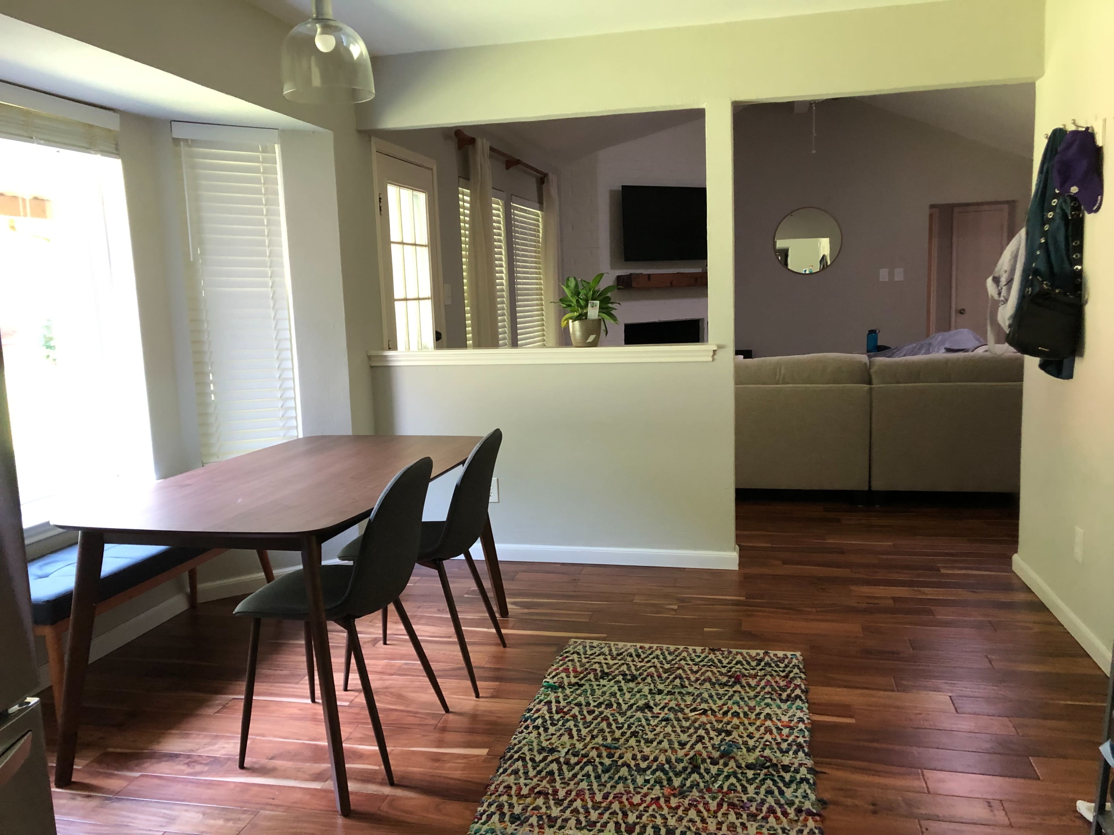A dining area with a wooden table and chairs, leading to a living room with a couch, and featuring large windows and natural wood flooring.