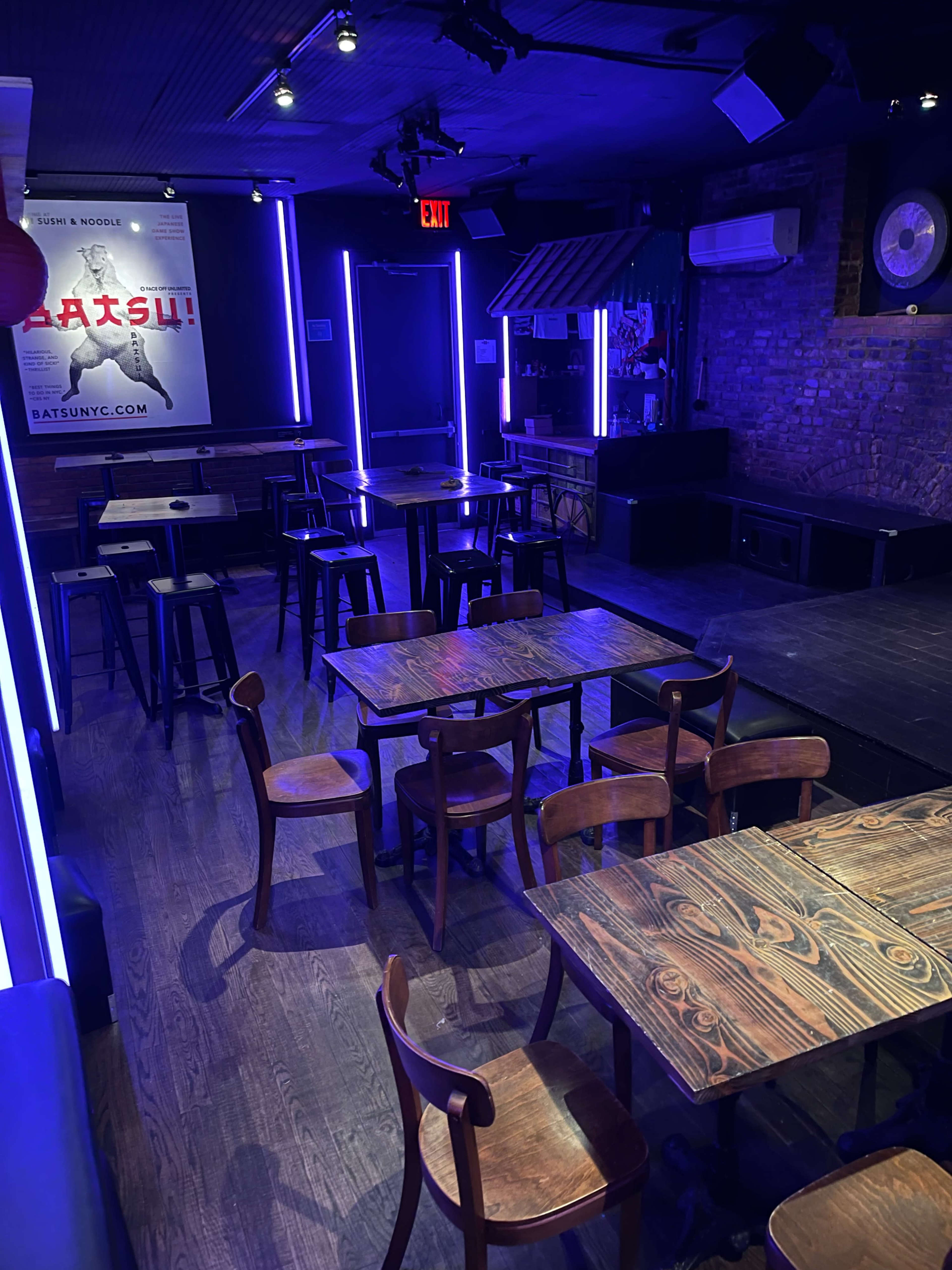 The image shows an empty music venue with wooden tables and chairs arranged in a dimly lit space featuring purple neon lighting.