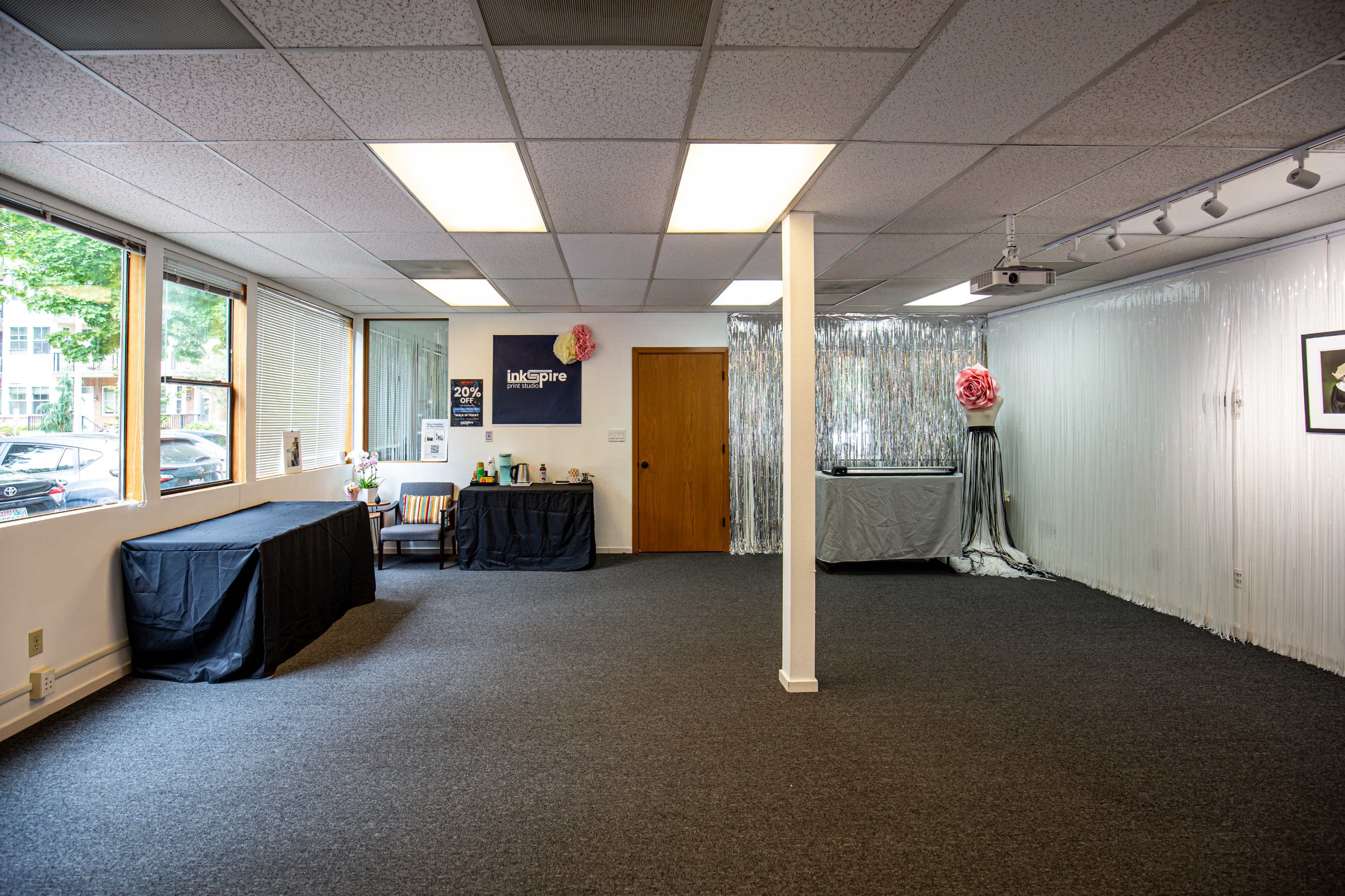 The image shows a spacious, empty room with grey carpet, large windows letting in natural light, and various decorative elements including a silver backdrop and tables covered with black cloth.