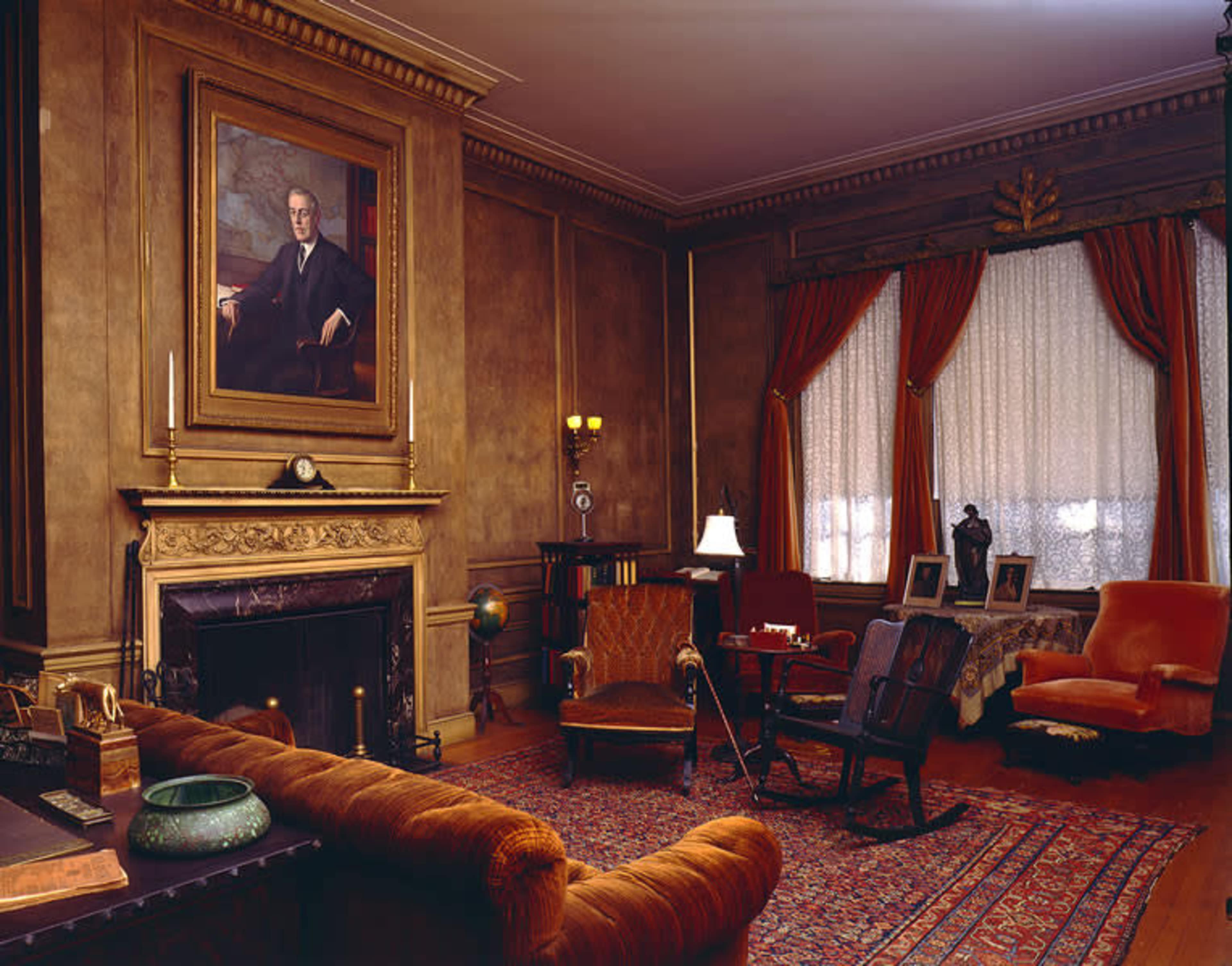 An elegantly furnished room featuring a prominent portrait above a fireplace, with antique furniture, a patterned rug, and warm lighting.