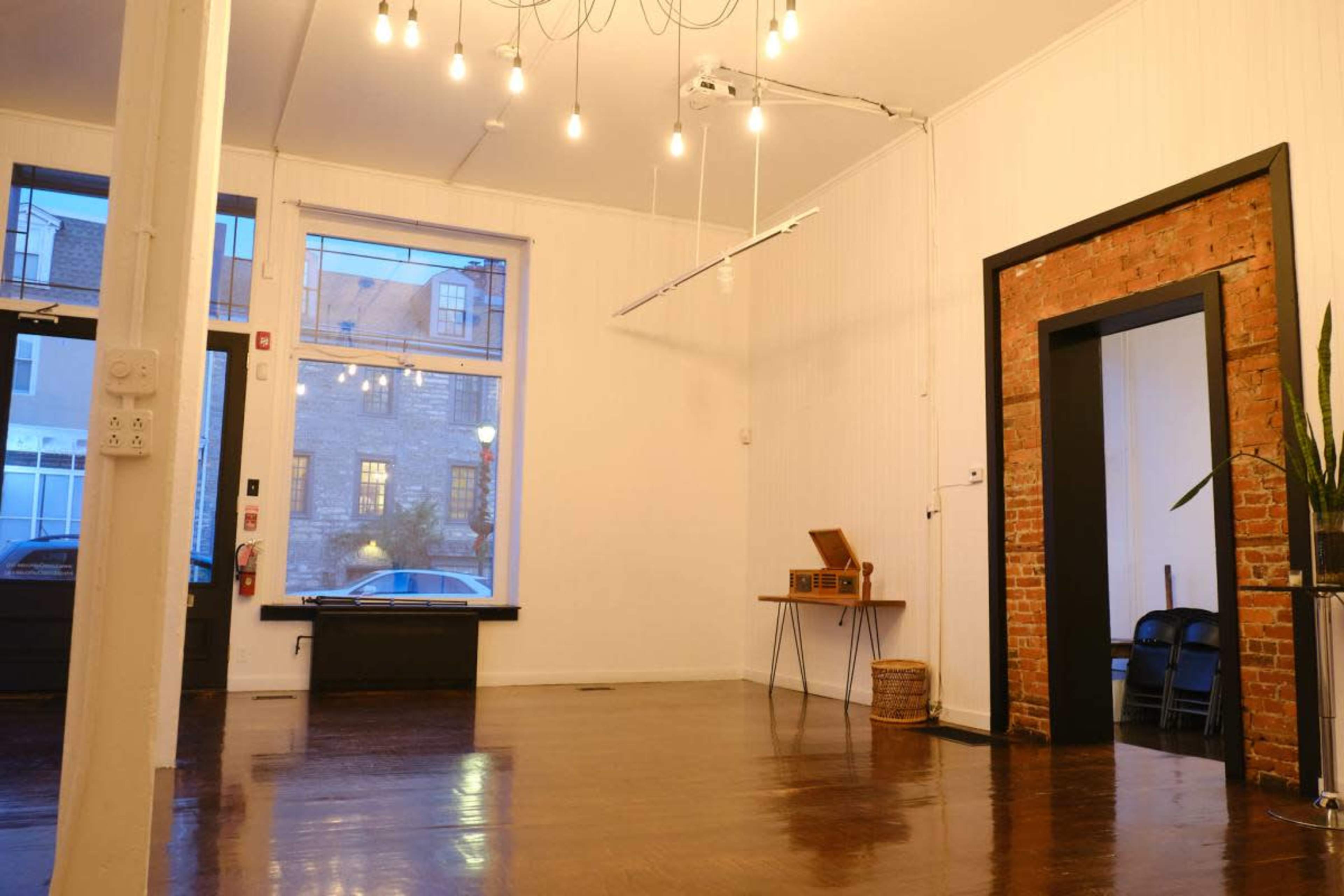The image shows a spacious indoor area with polished wooden floors, natural light from a large window, and exposed brick elements on the walls.