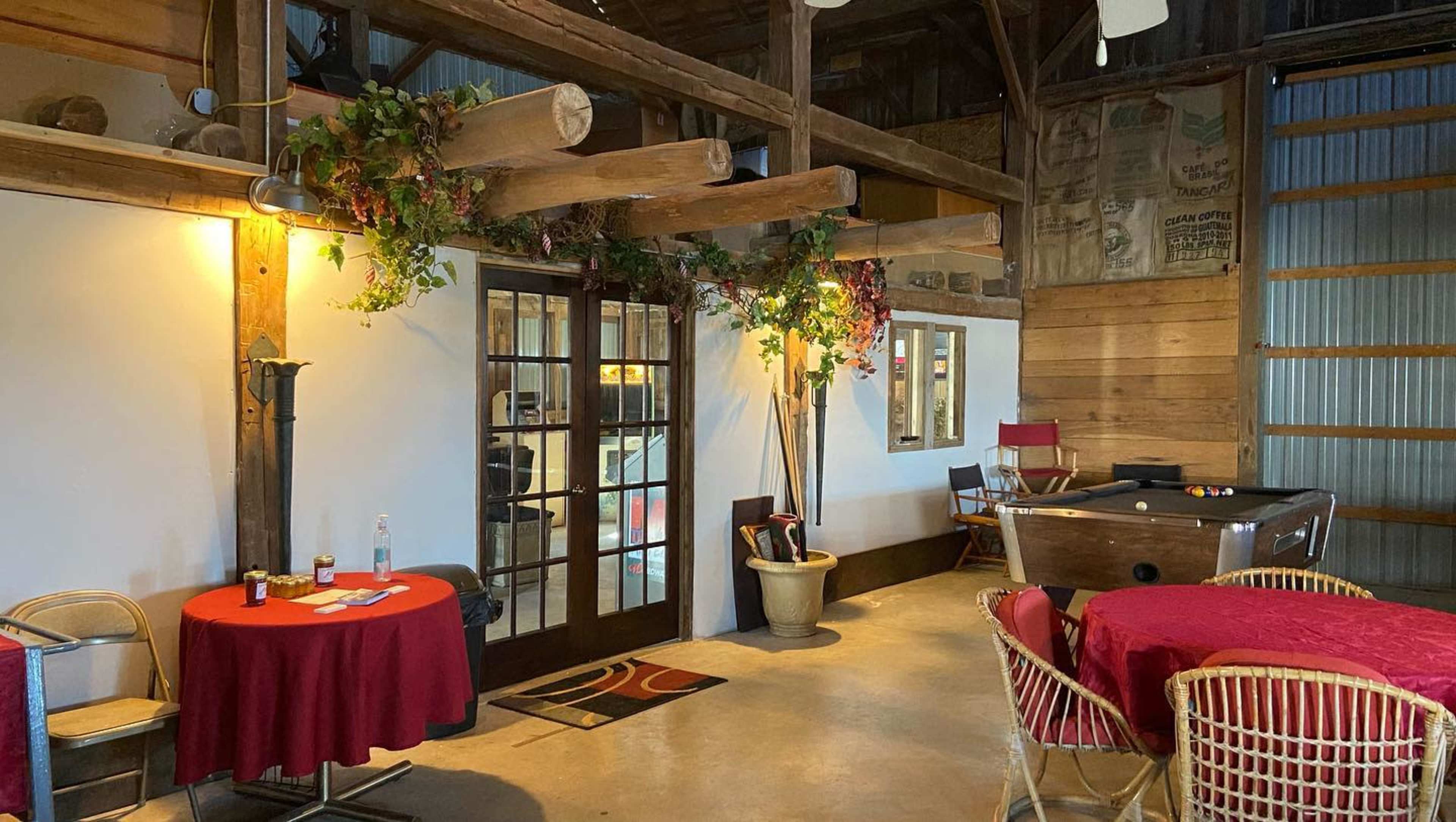 The interior of a rustic room features wooden beams, tables set with red linens, and a pool table in the corner.