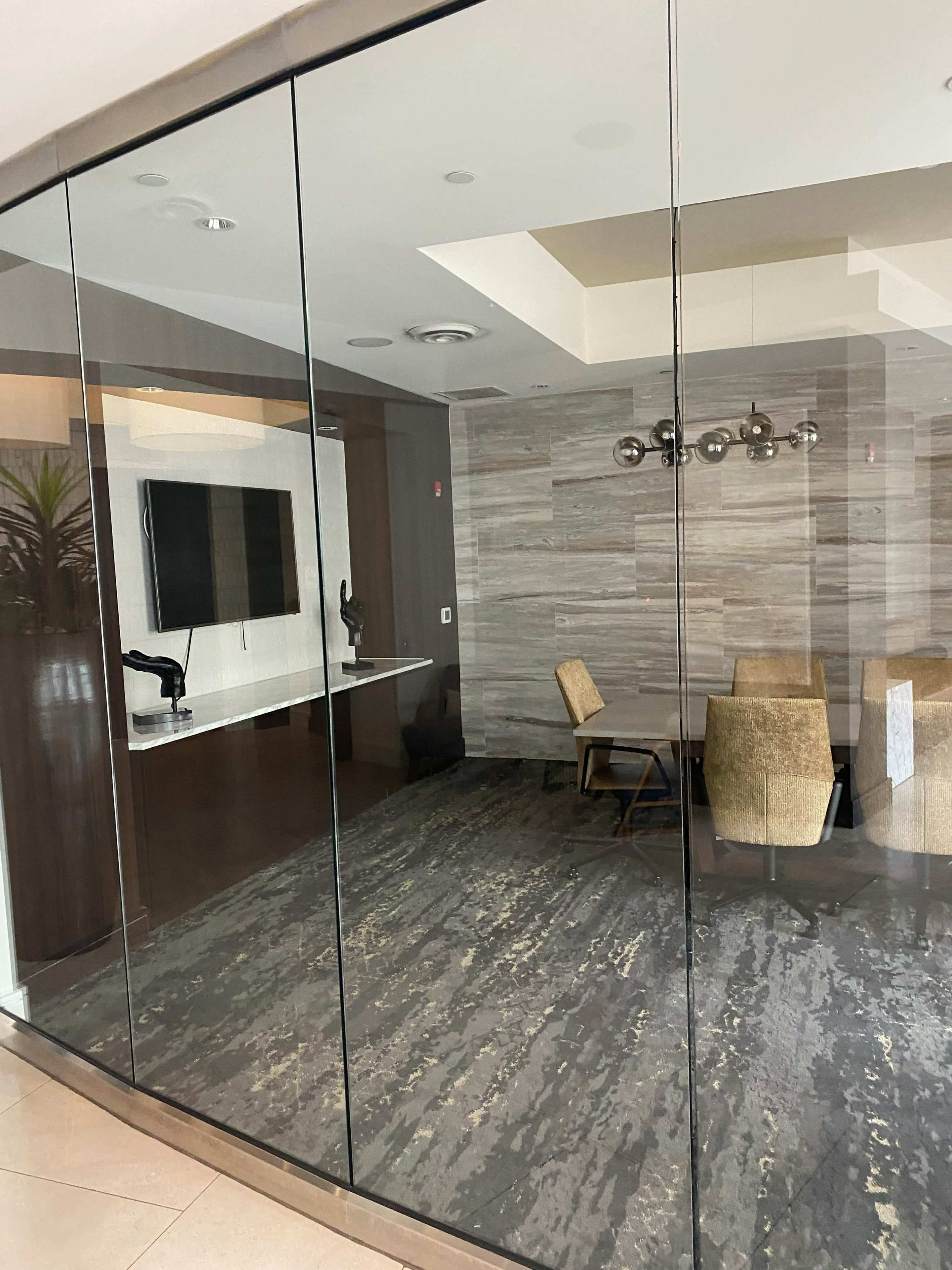 A modern conference room enclosed by glass walls, featuring a table surrounded by chairs and a wall-mounted screen.
