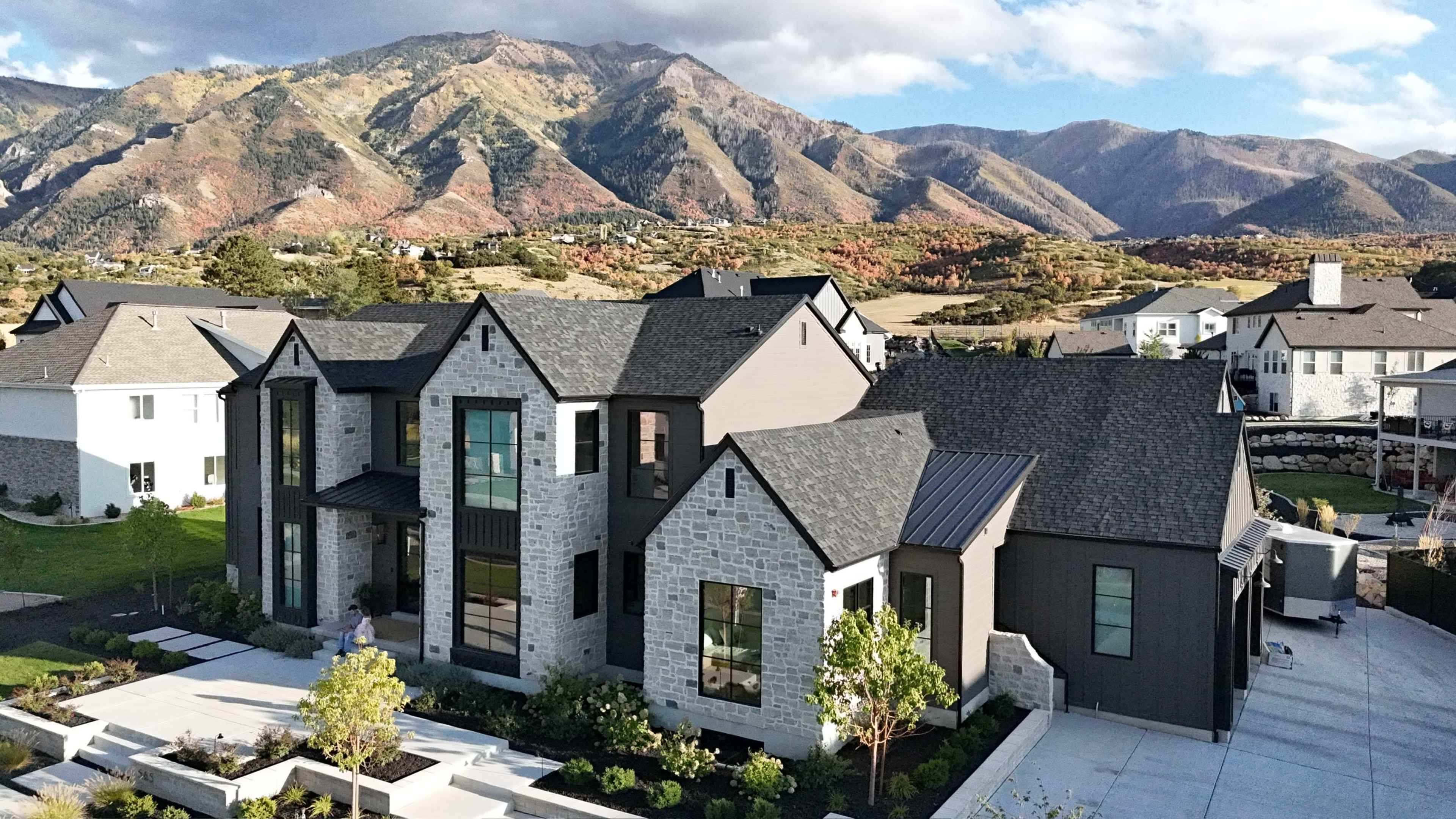 A large, modern home with a stone and dark exterior is set against a backdrop of rolling mountains and a clear sky.