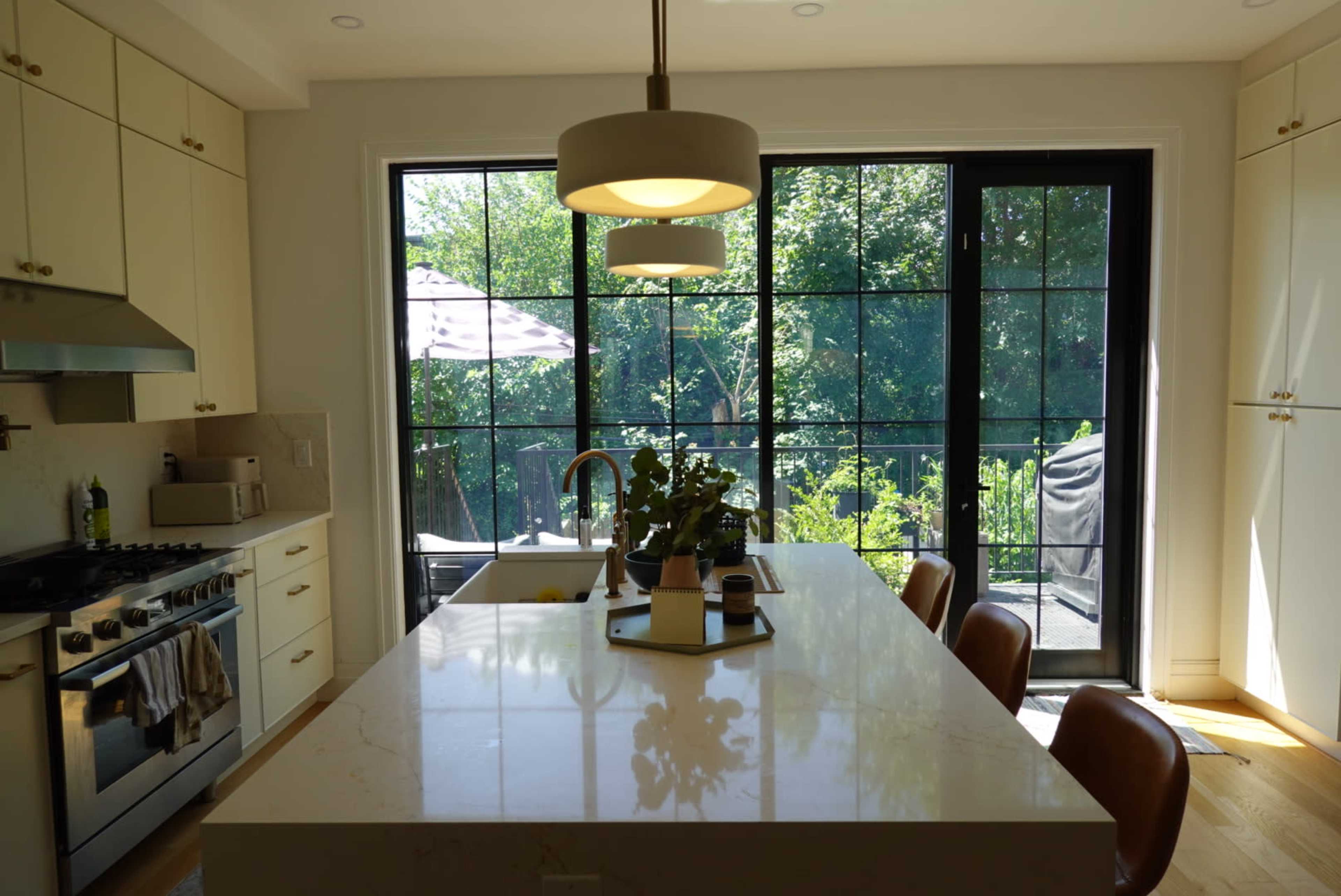 The image depicts a modern kitchen featuring a central island with seating, large windows overlooking a garden, and a stainless steel stove.
