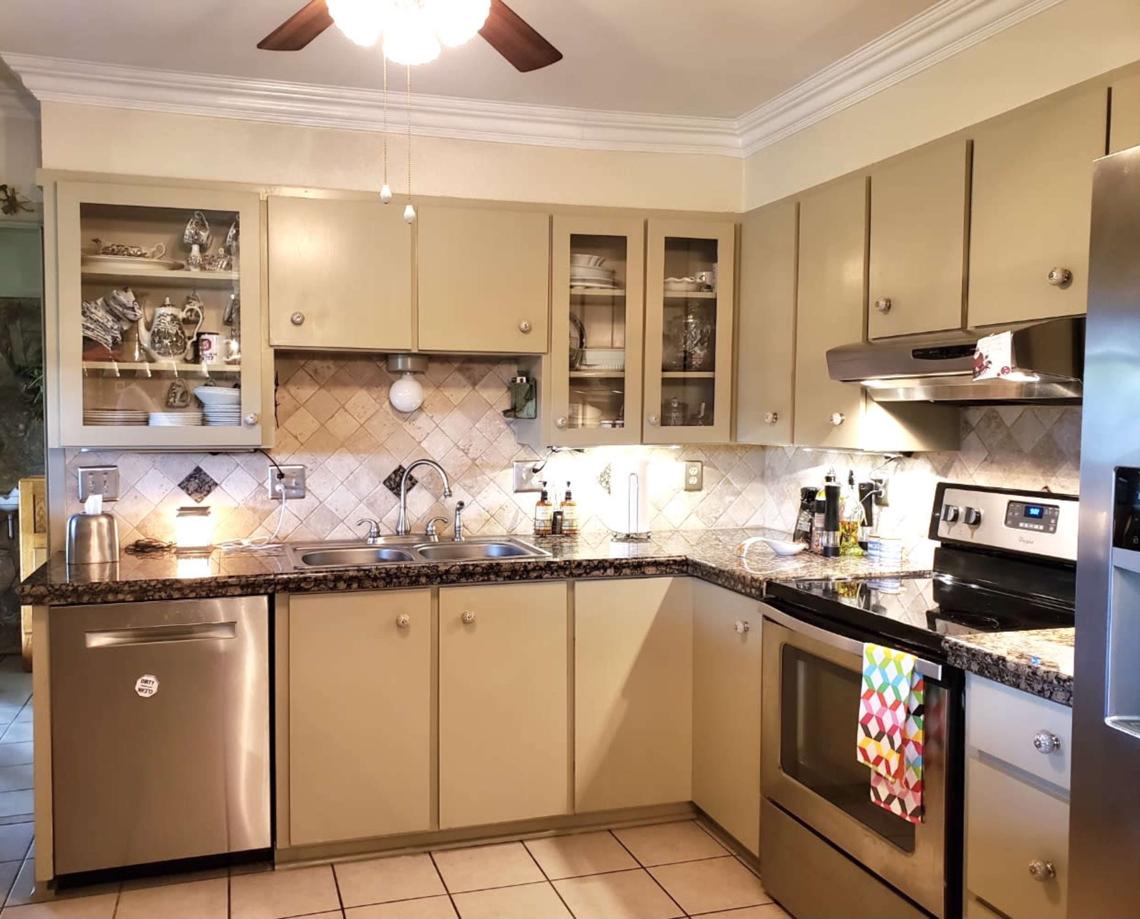 The image shows a kitchen with beige cabinets, stainless steel appliances, and a tiled backsplash, featuring a sink and organized dishware in glass-fronted cabinets.