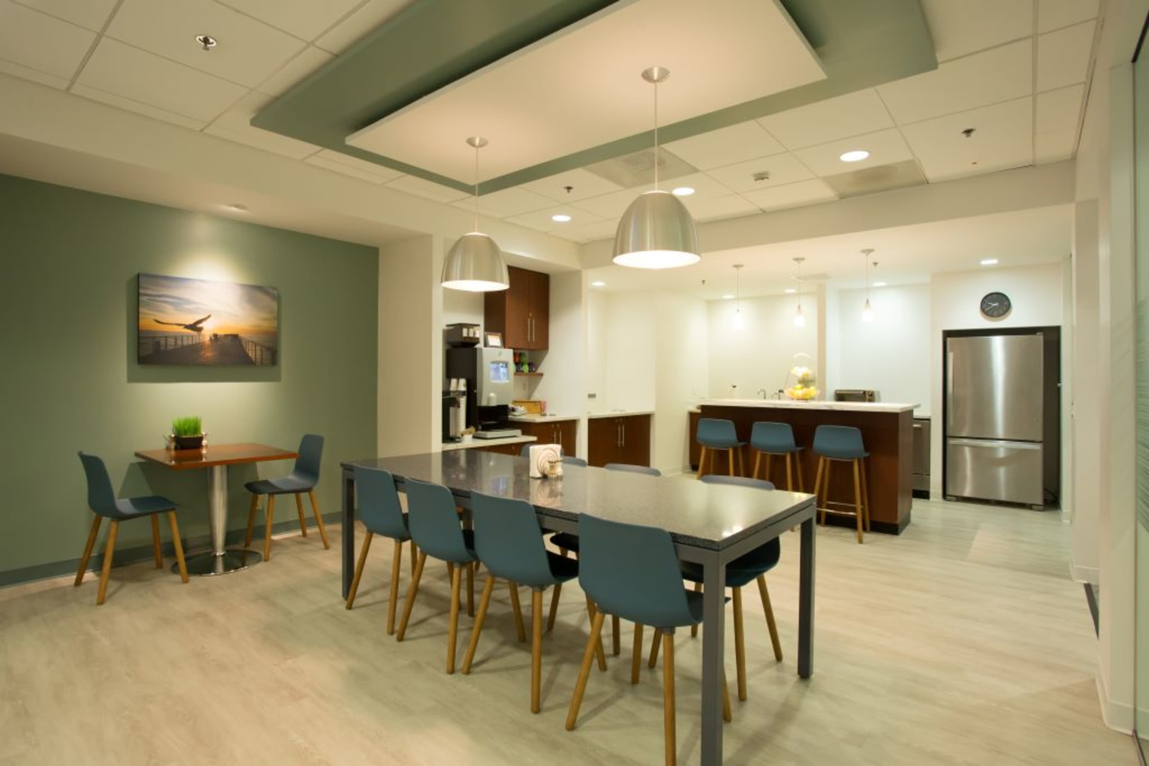 A modern kitchen and dining area features a large table with chairs, a bar counter, and kitchen appliances against a light-colored backdrop.