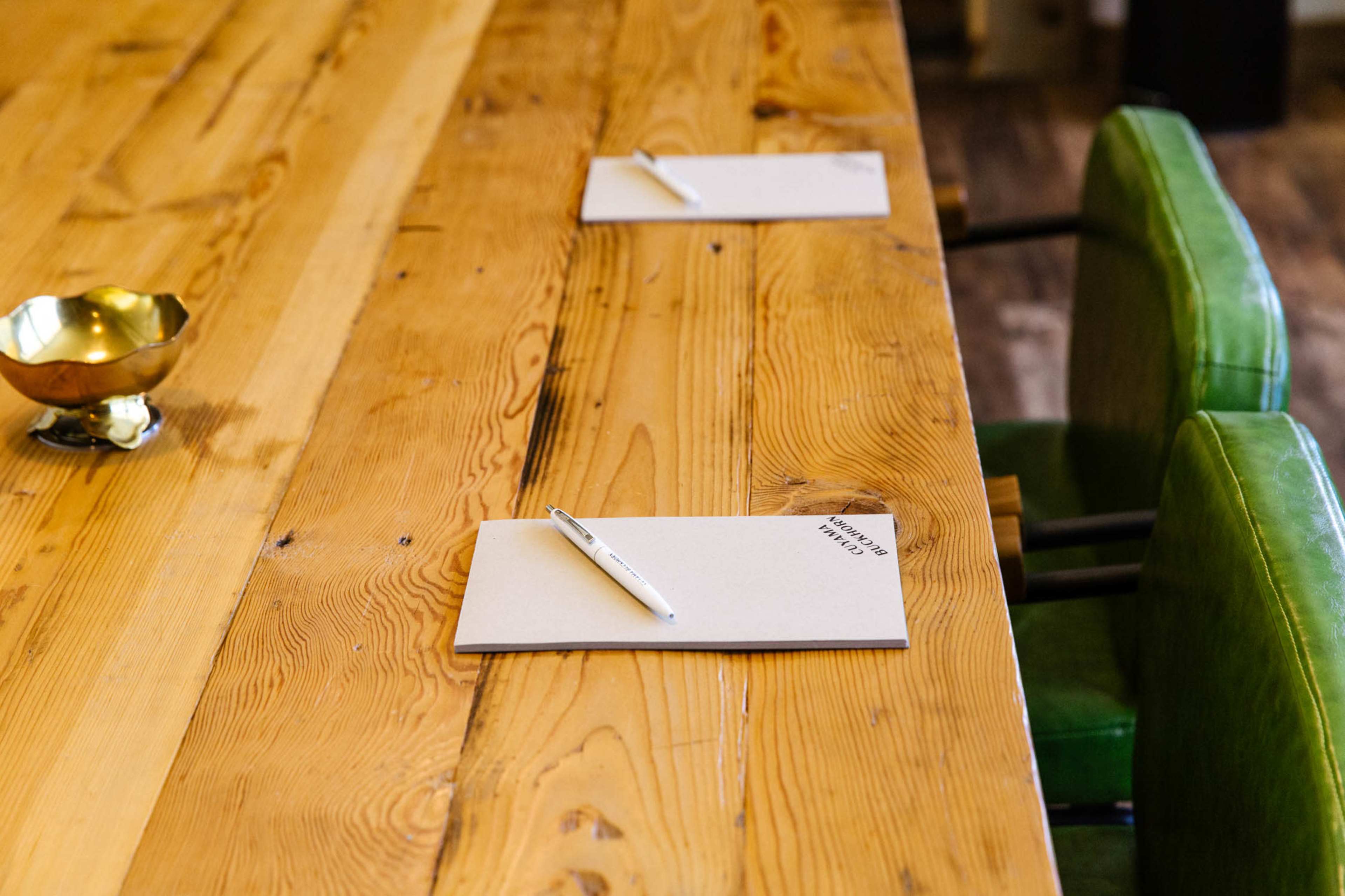 A wooden table with two notepads and pens placed on it, alongside a small bowl, and two green chairs positioned at the sides.