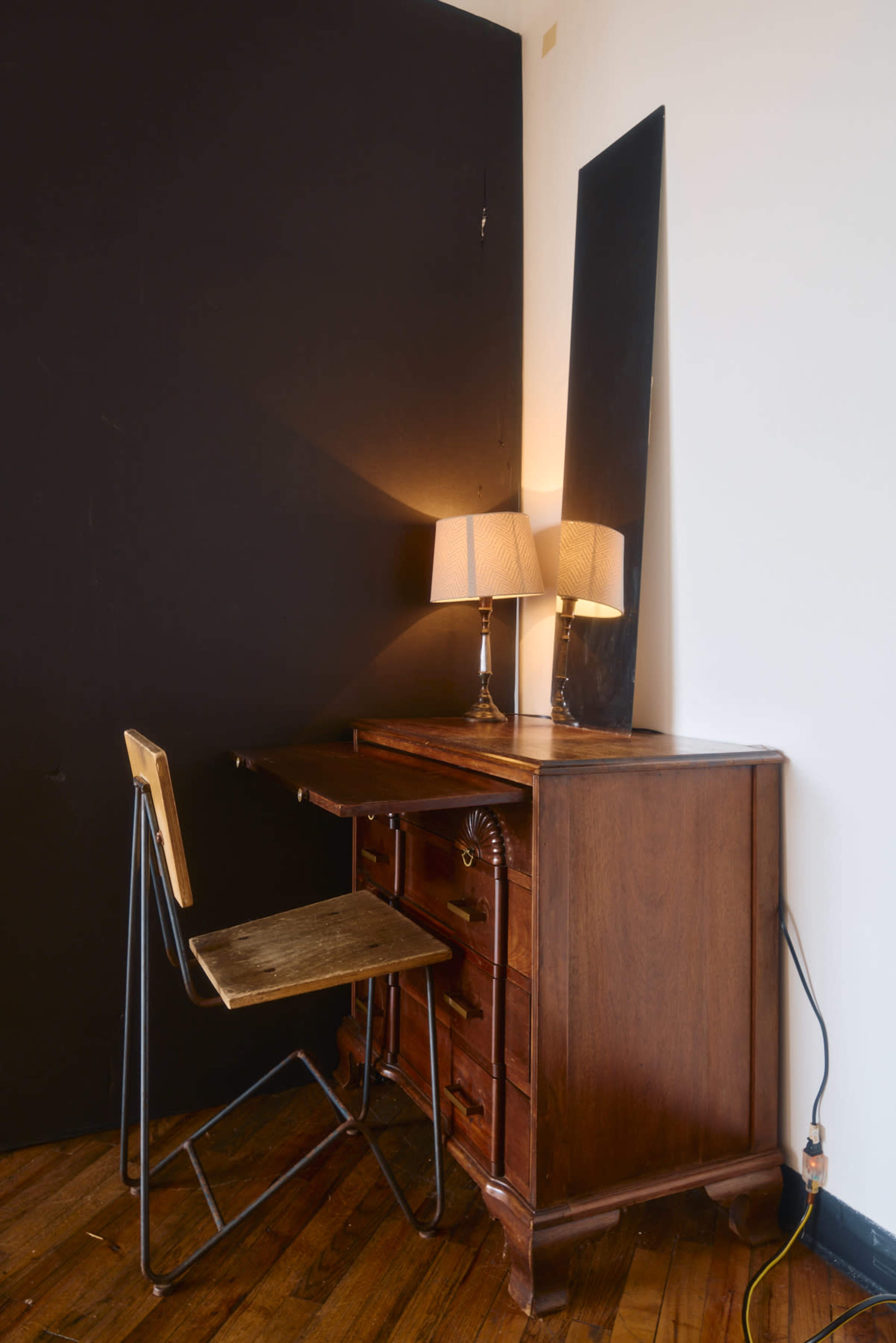 A wooden writing desk with drawers is positioned against a black wall, accompanied by a folding chair and two lamps providing soft lighting.