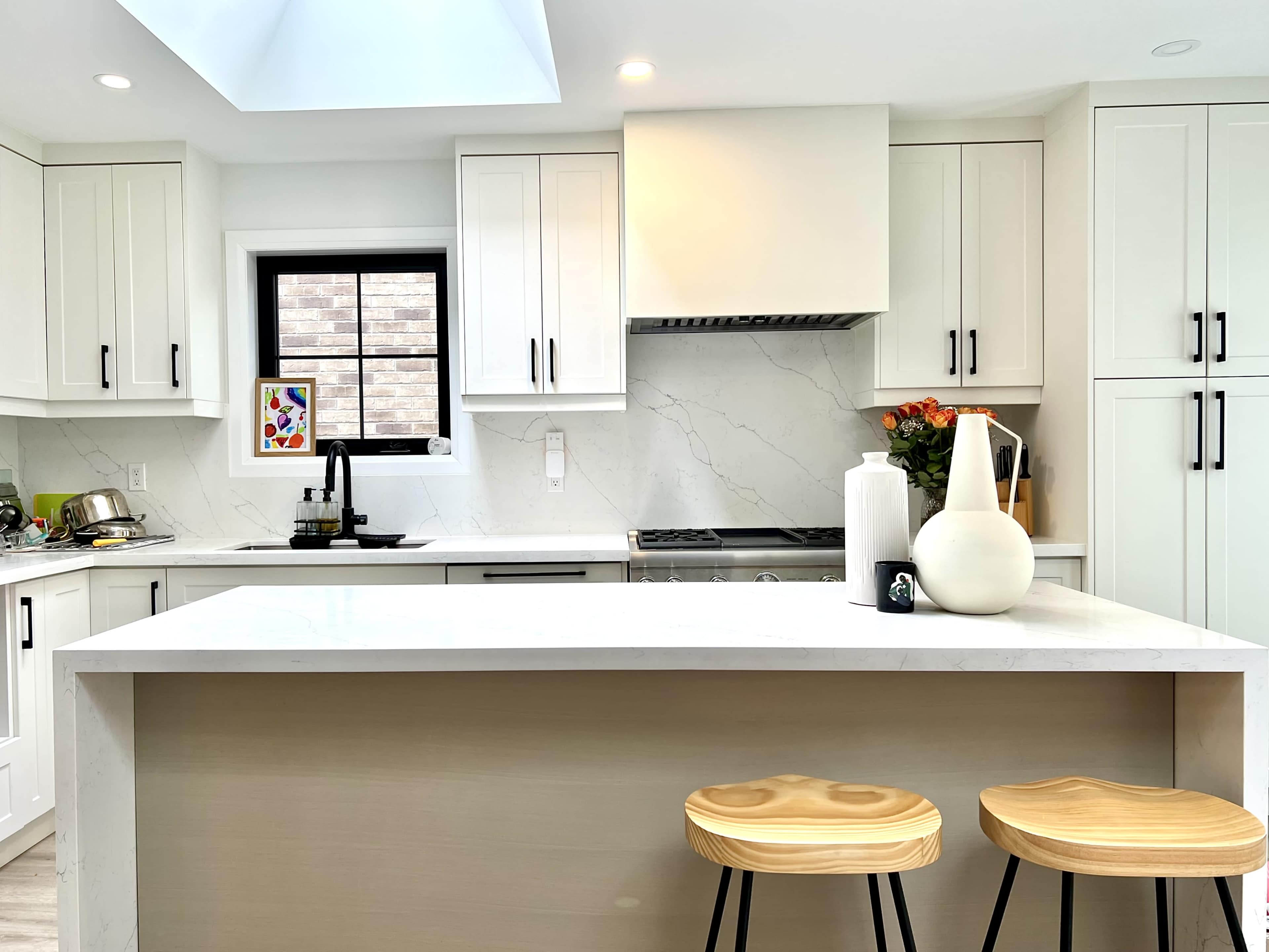 The image shows a modern kitchen with white cabinets, a marble countertop, and two wooden stools at the island.