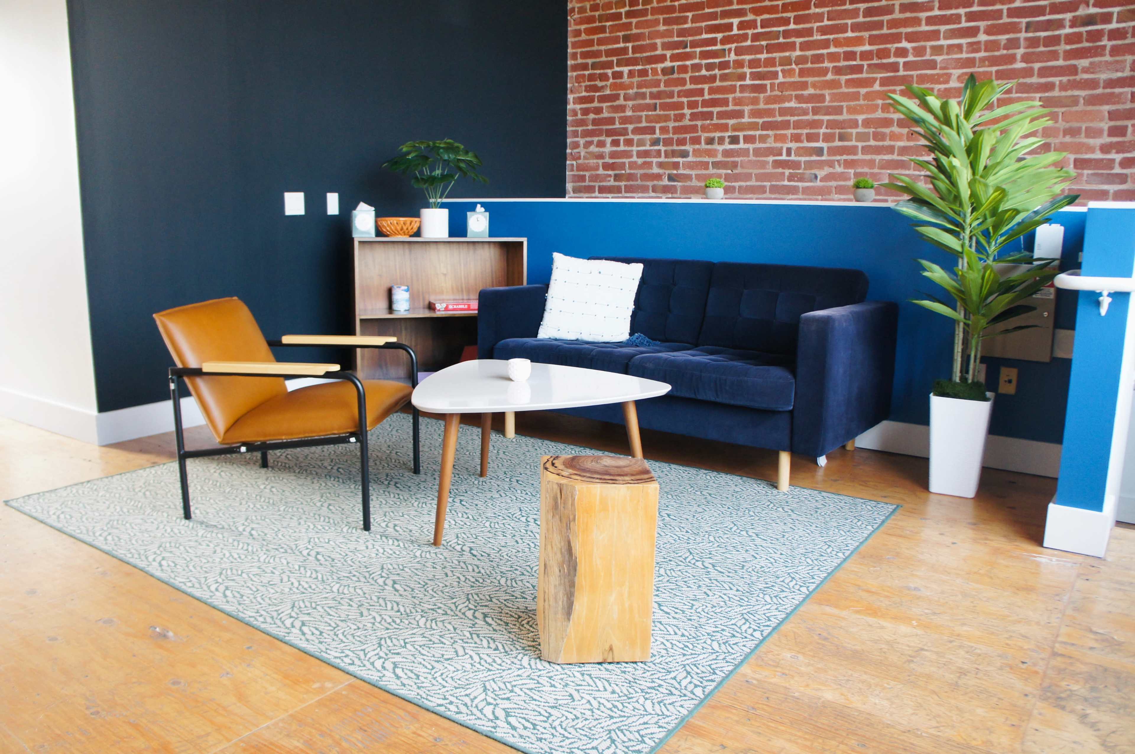 The image shows a modern living room with a navy blue sofa, an orange accent chair, a round coffee table, a wooden stool, and a green area rug on a wooden floor, set against a backdrop of exposed brick and blue walls.
