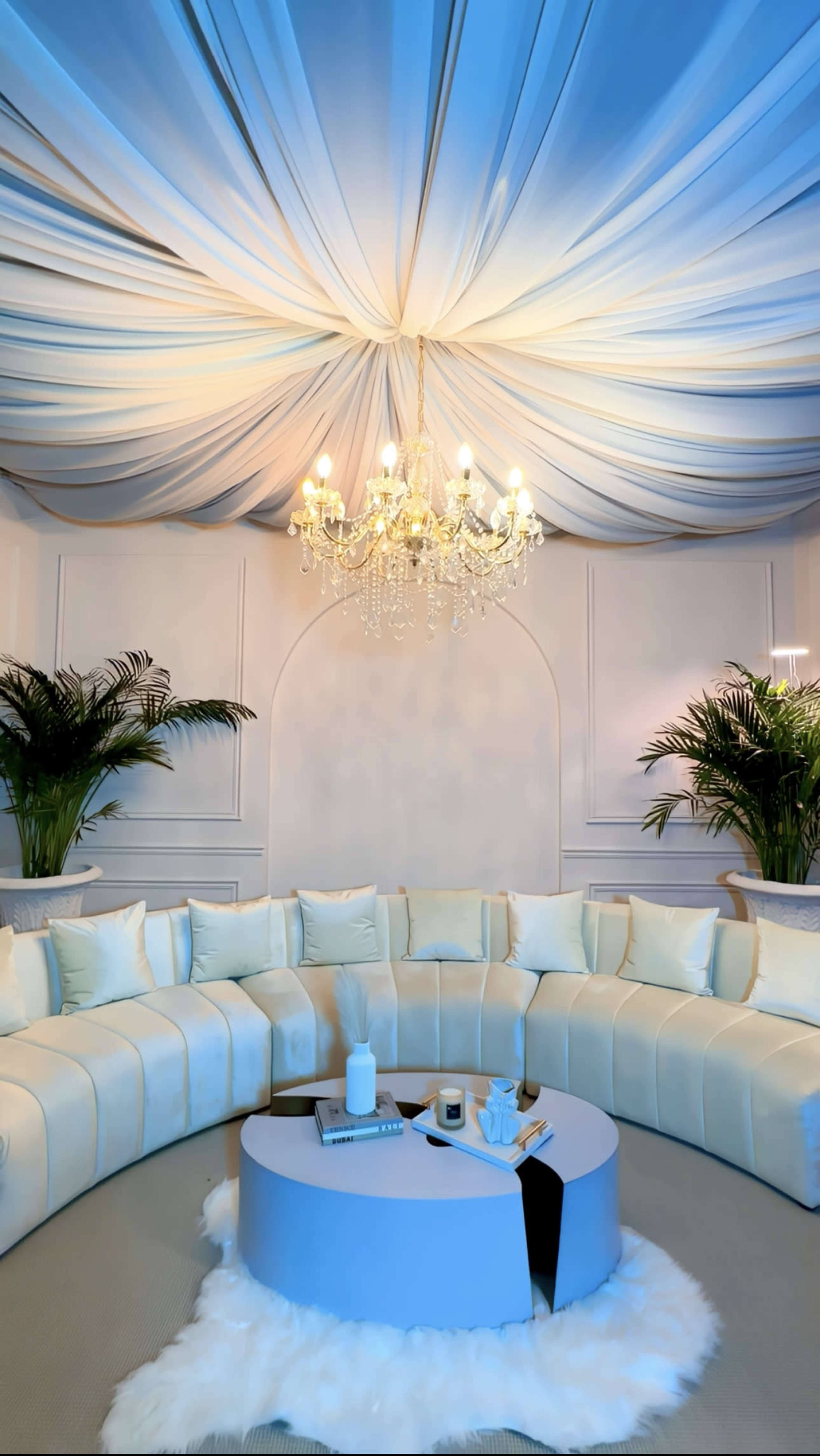 A spacious lounge area features a large white sectional couch, a circular coffee table, and decorative plants under a draped ceiling with a chandelier.