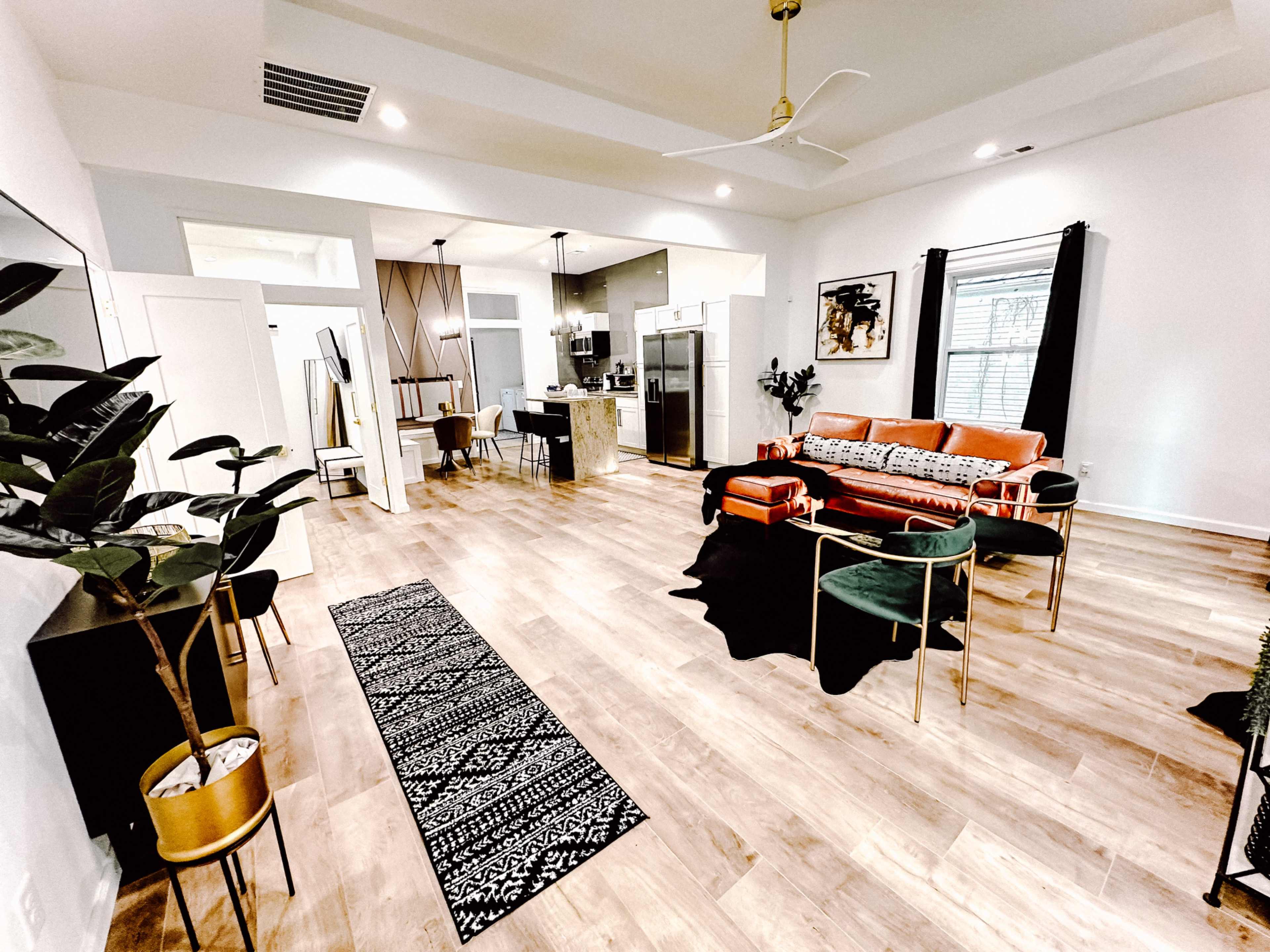 The spacious living area features a bright, open layout with a combination of modern furniture, including a coral sofa, plants, and a patterned area rug.