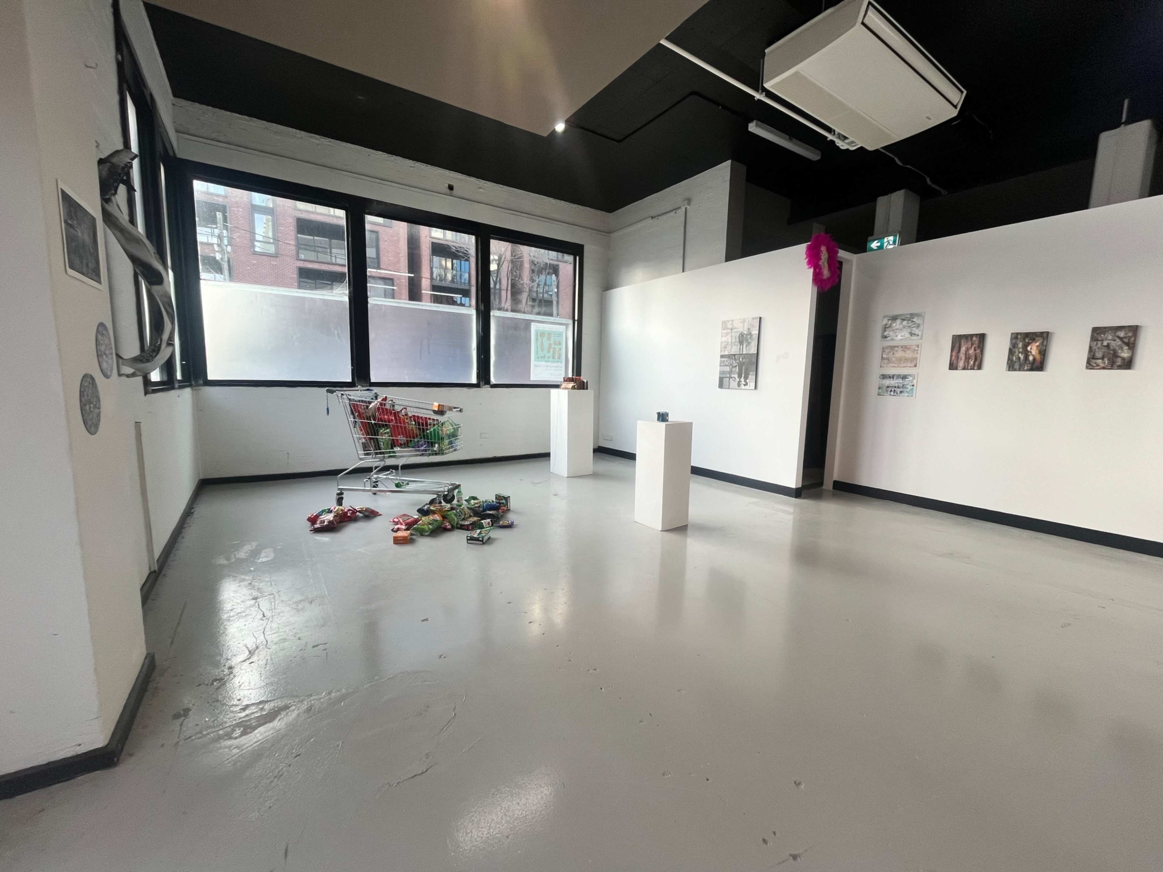 The image shows an art gallery interior with large windows, white walls displaying various artworks, and a shopping cart filled with colorful items placed on the floor.