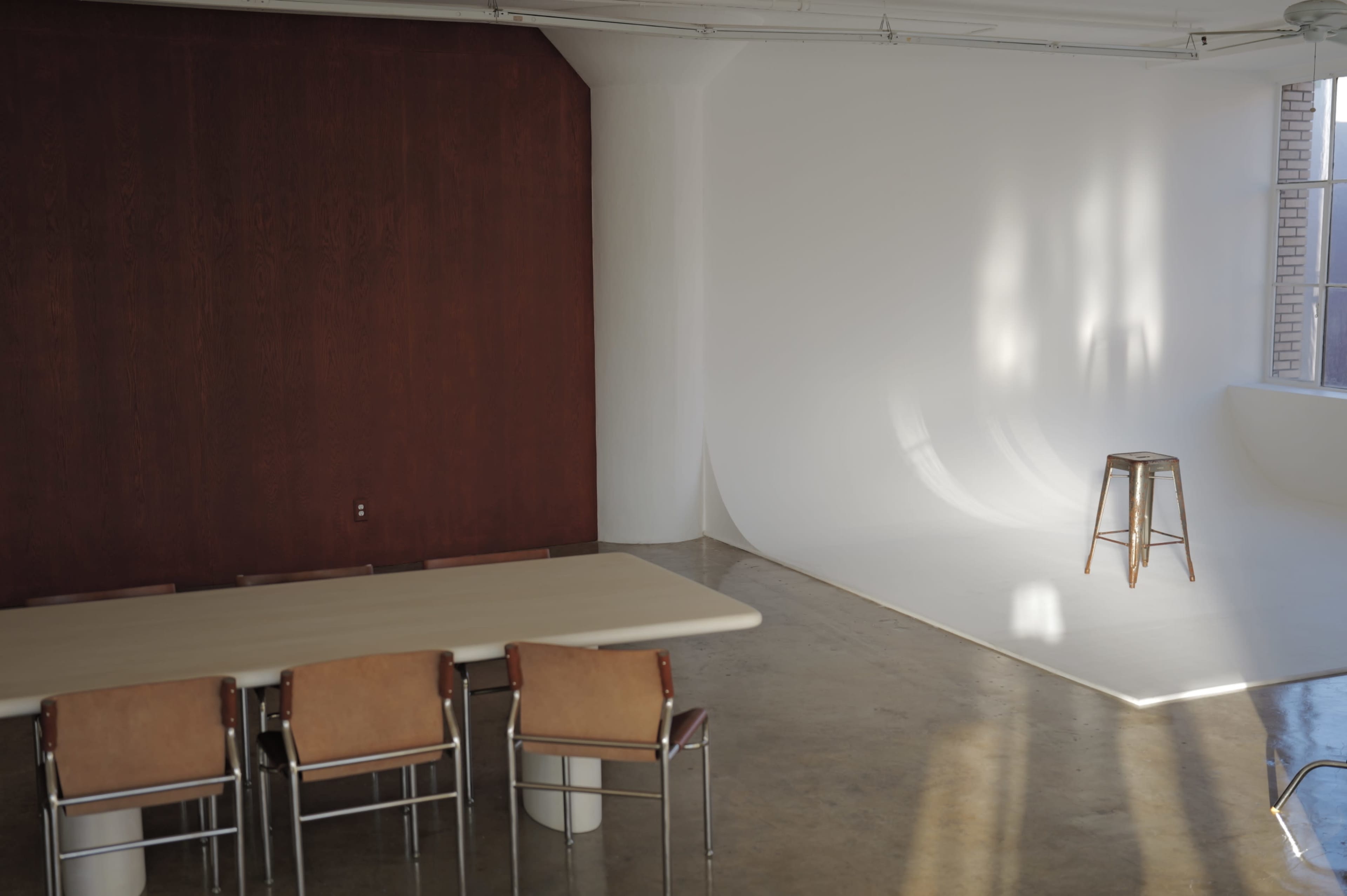 The image shows a minimalist interior with a wooden wall, a light-colored table, and several chairs arranged around it, alongside a simple backdrop and a wooden stool in the corner.