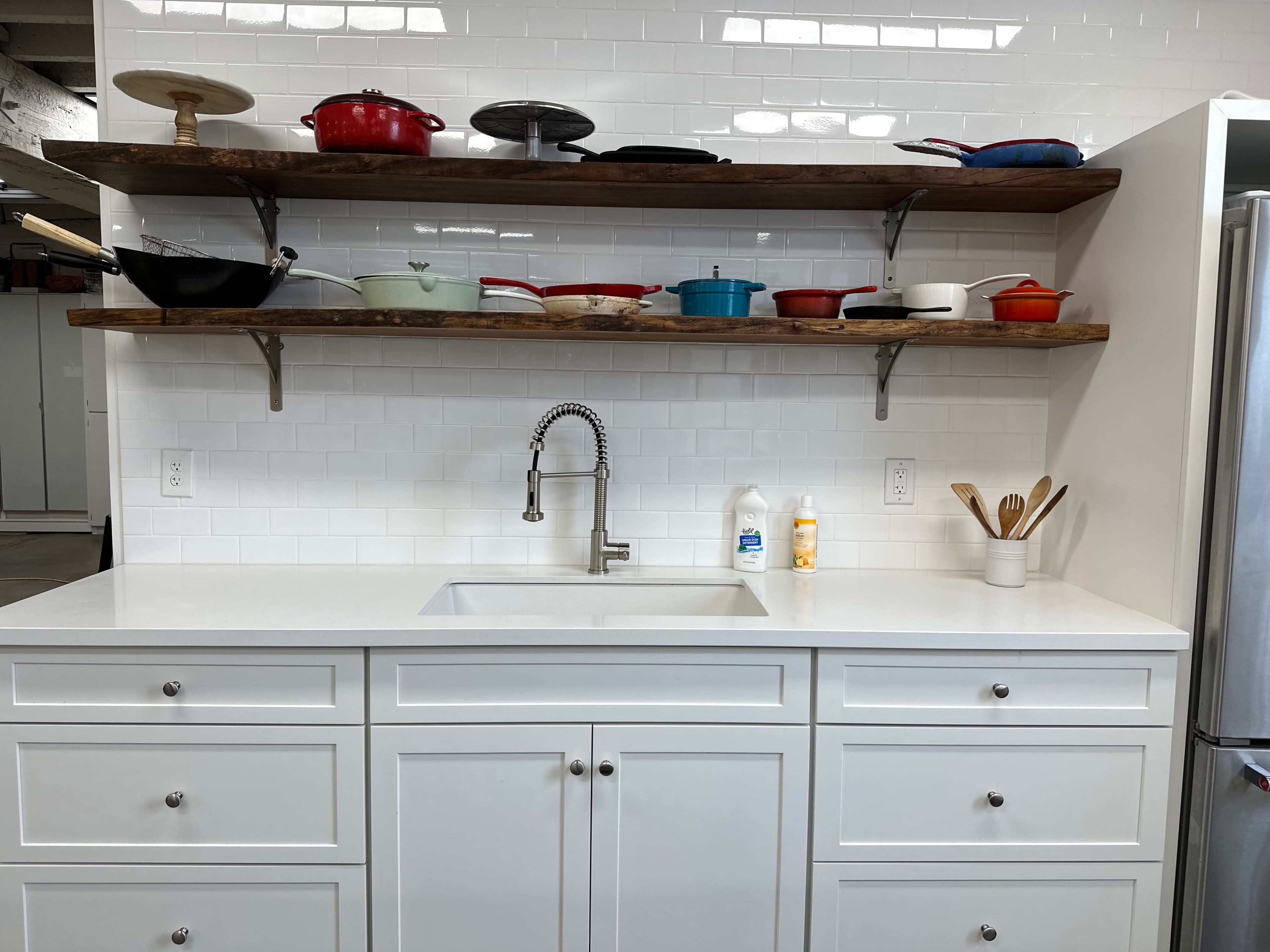 A modern kitchen features a white countertop and cabinetry, with open wooden shelves displaying various cookware above.