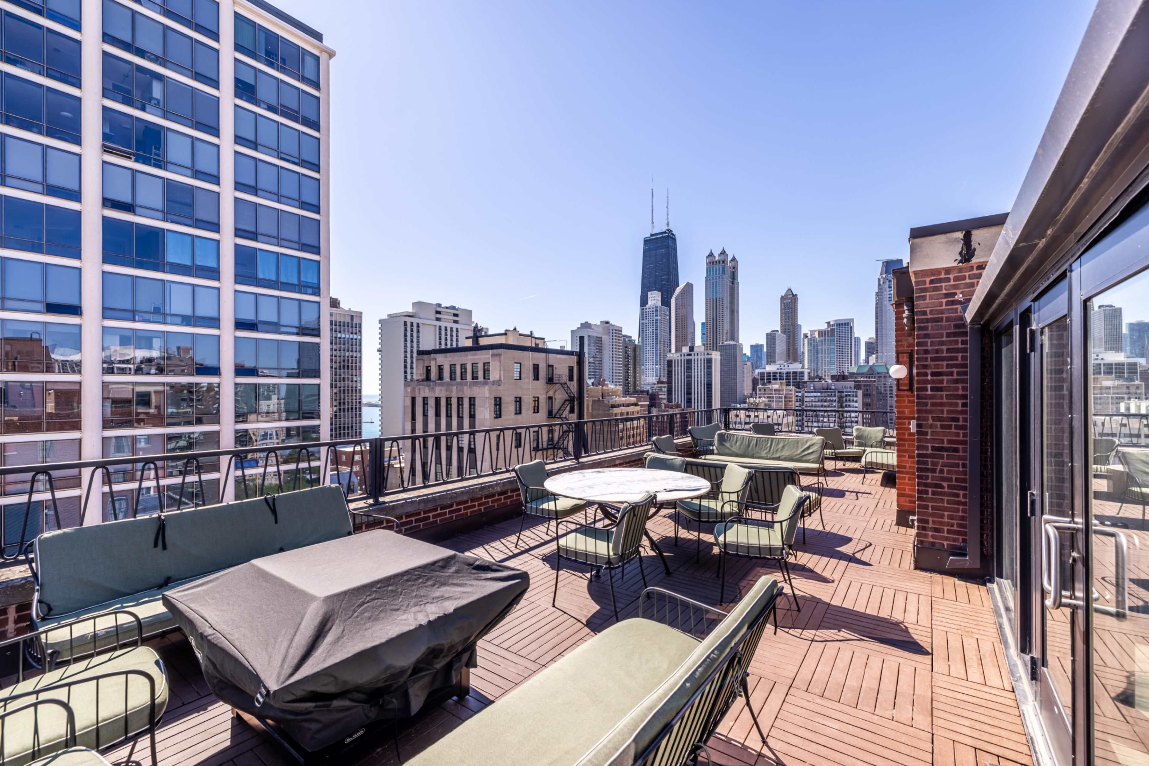 The image shows a rooftop terrace with seating areas and tables, overlooking a city skyline featuring tall buildings in clear weather.
