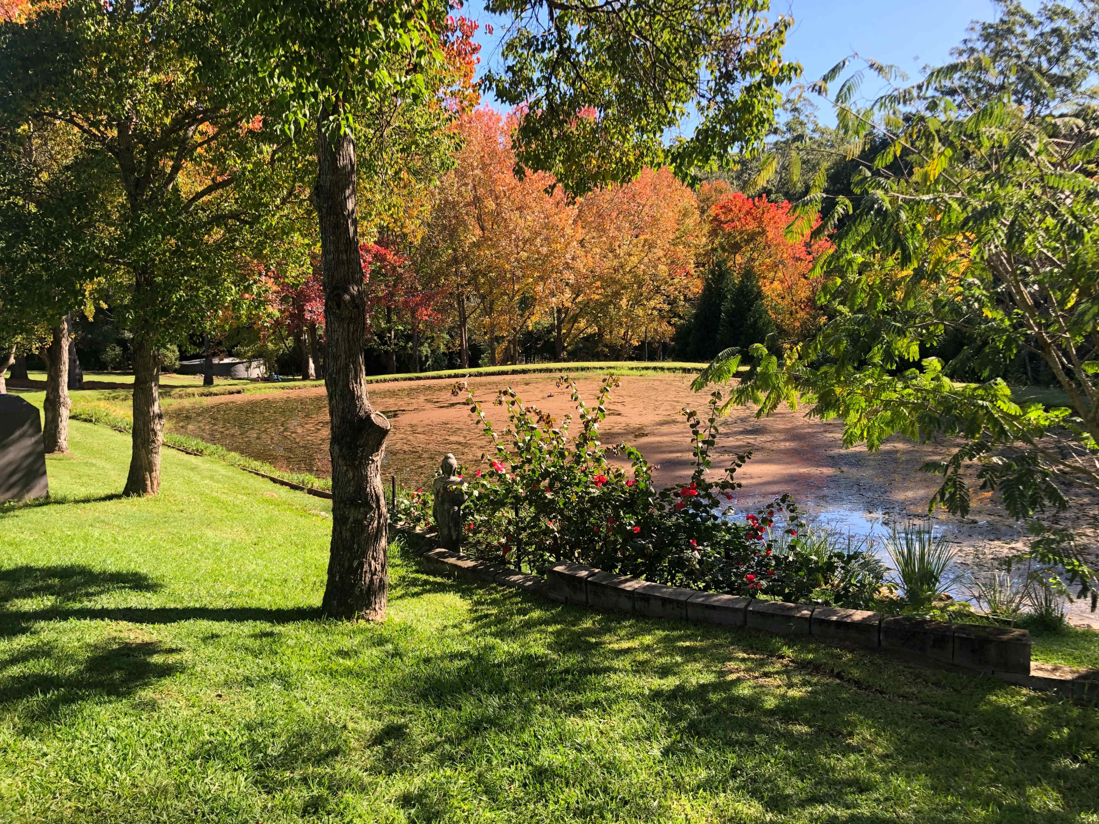 A pond is surrounded by trees and colorful autumn foliage with a well-maintained grassy area and flower beds in the foreground.