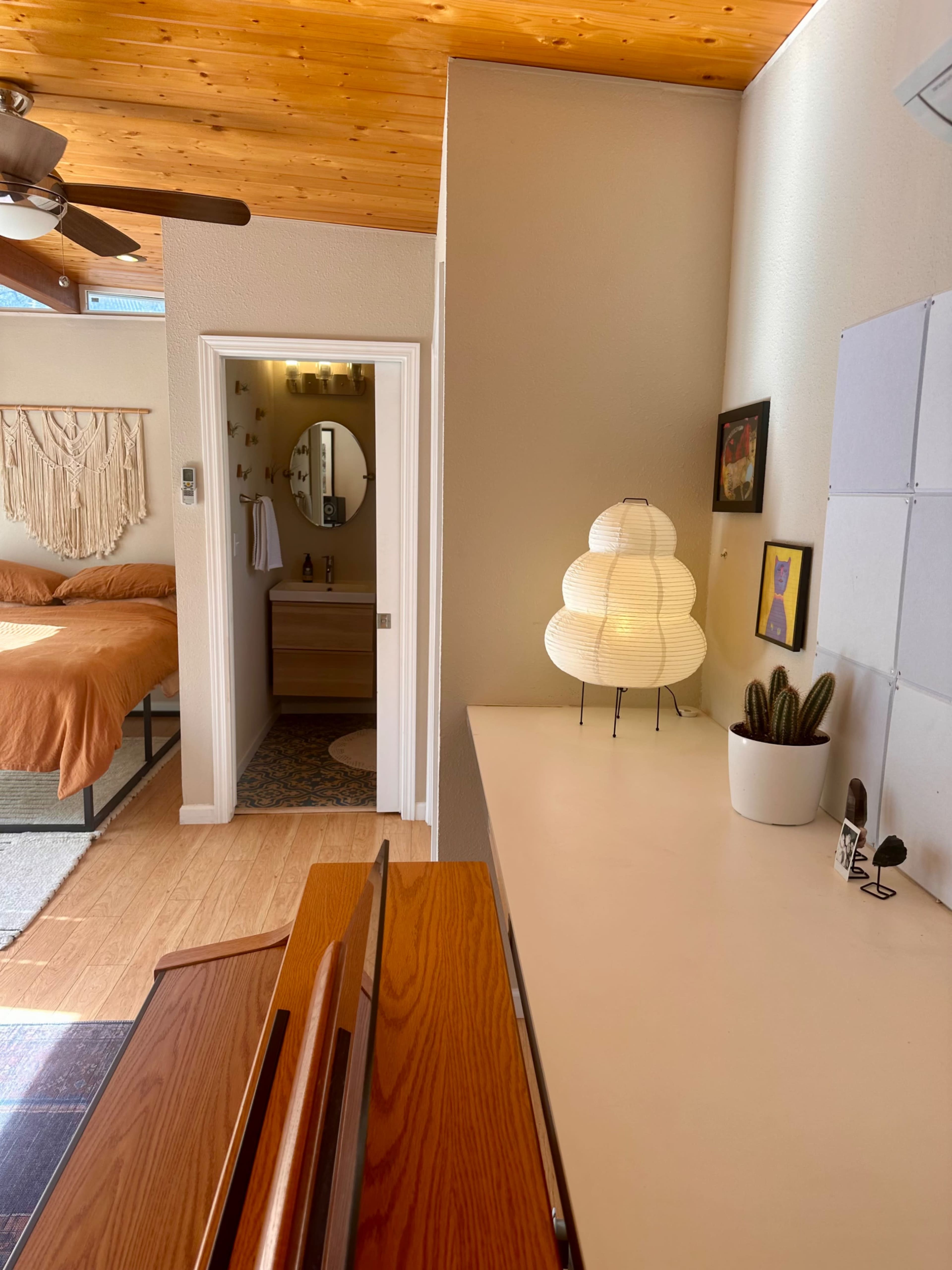 The image shows a bright room with a wooden ceiling, featuring a bed in the background, a round mirror, a light fixture on a console table, and a small plant on display.