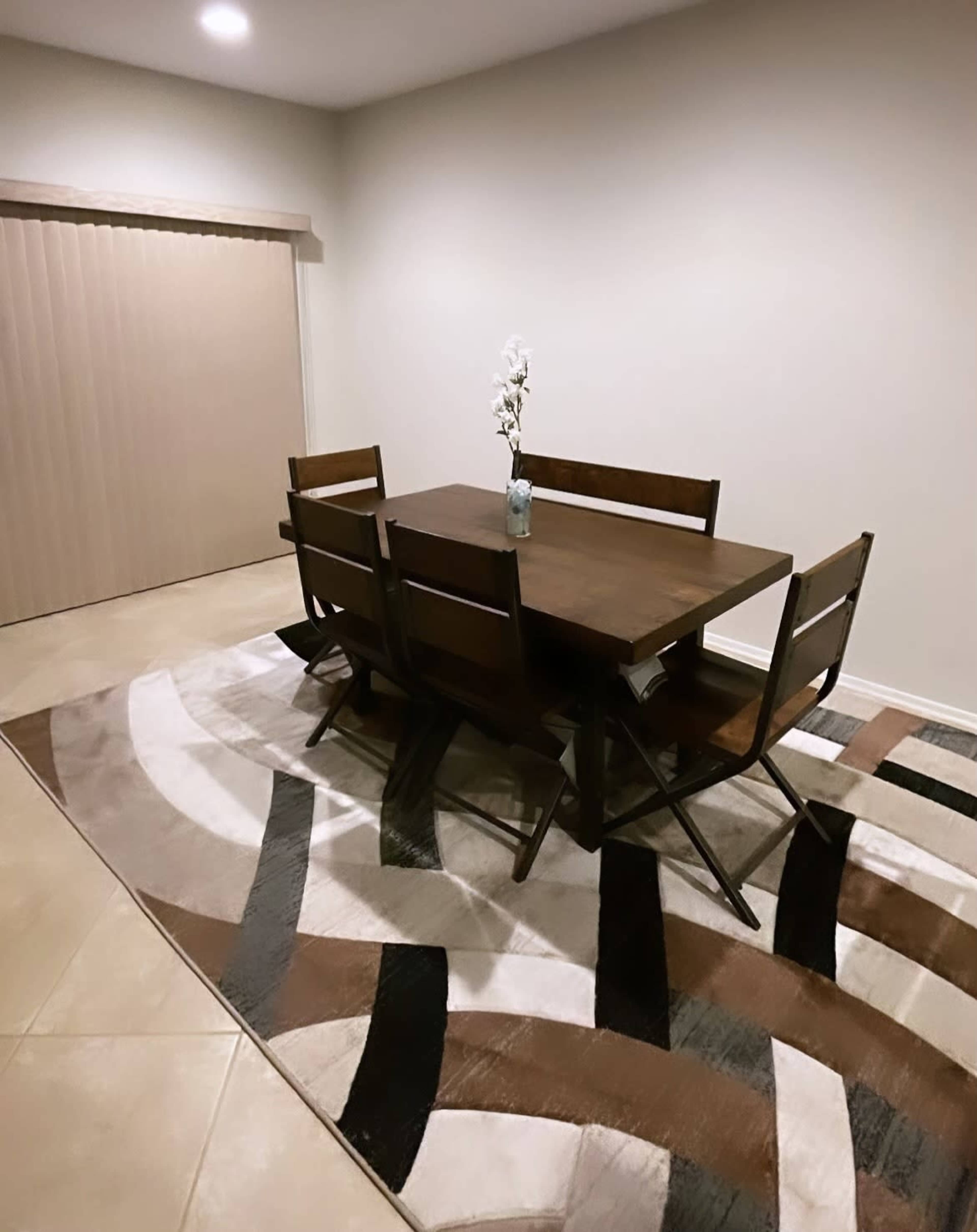 A wooden dining table with four chairs is set on a patterned rug in a well-lit room with a sliding door.