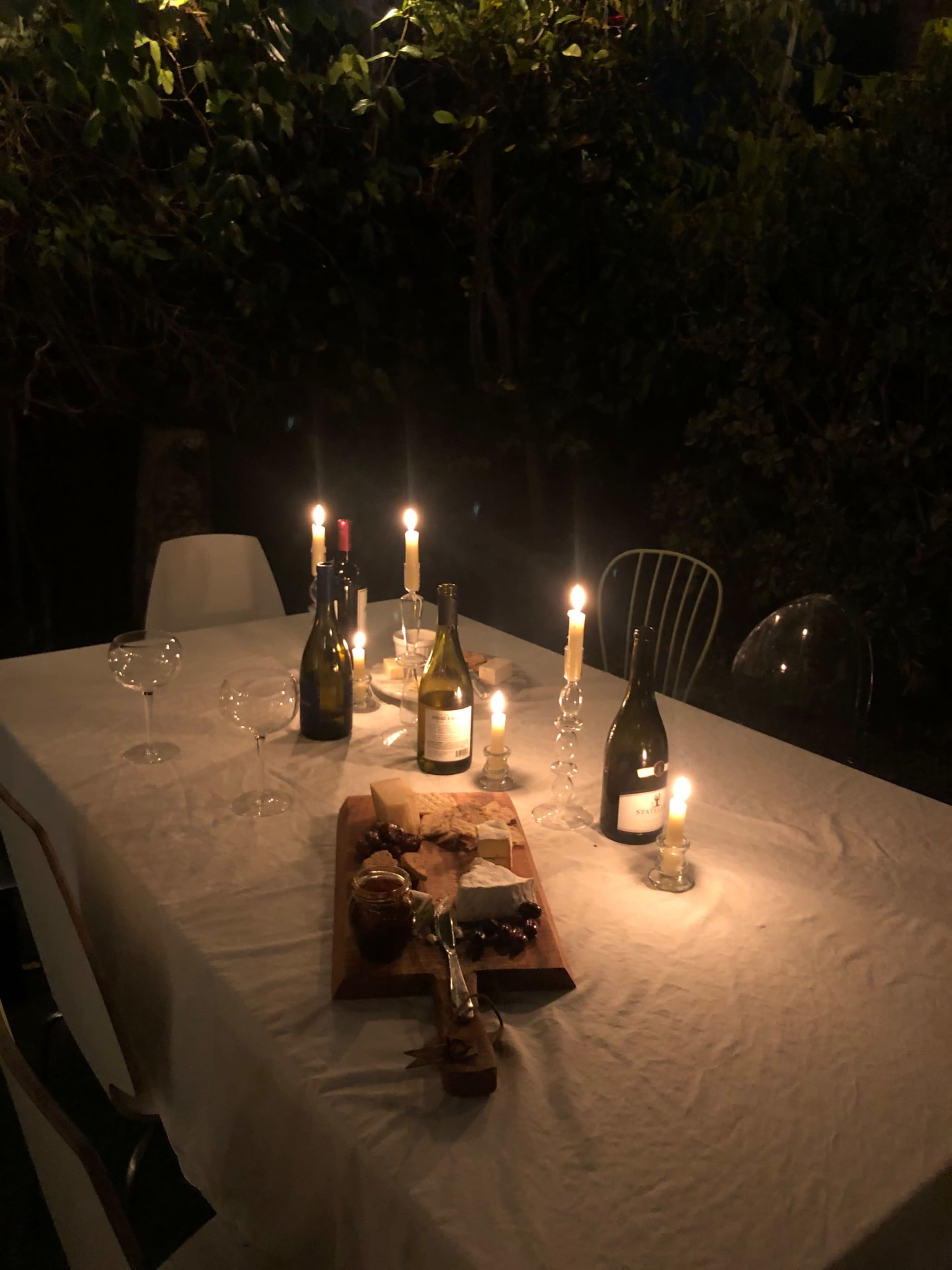 A table set outdoors is adorned with candles, wine bottles, and a wooden platter of food in a softly lit atmosphere.