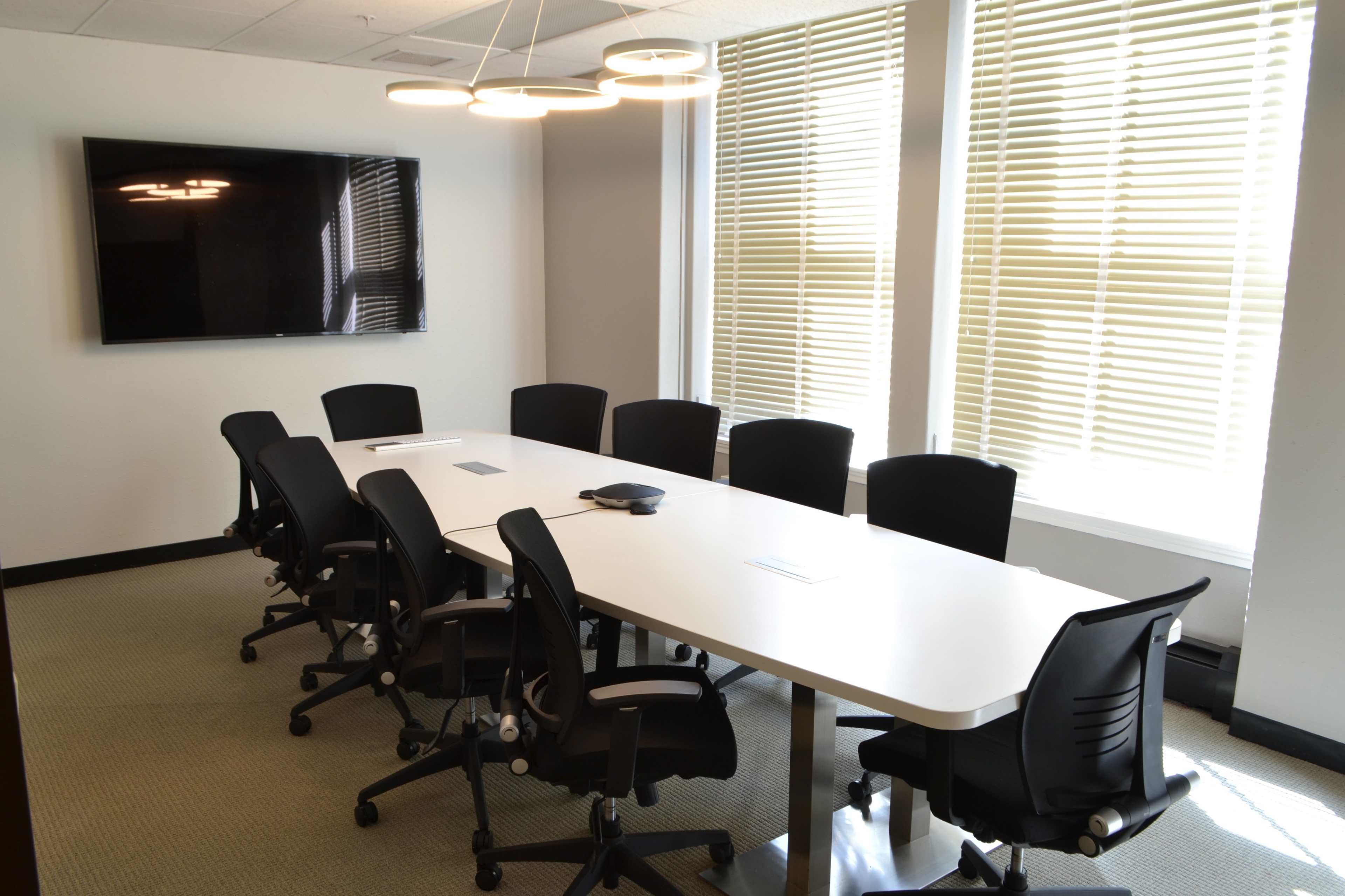 A modern conference room features a long white table surrounded by black ergonomic chairs, with a large TV on the wall and natural light filtering through window blinds.