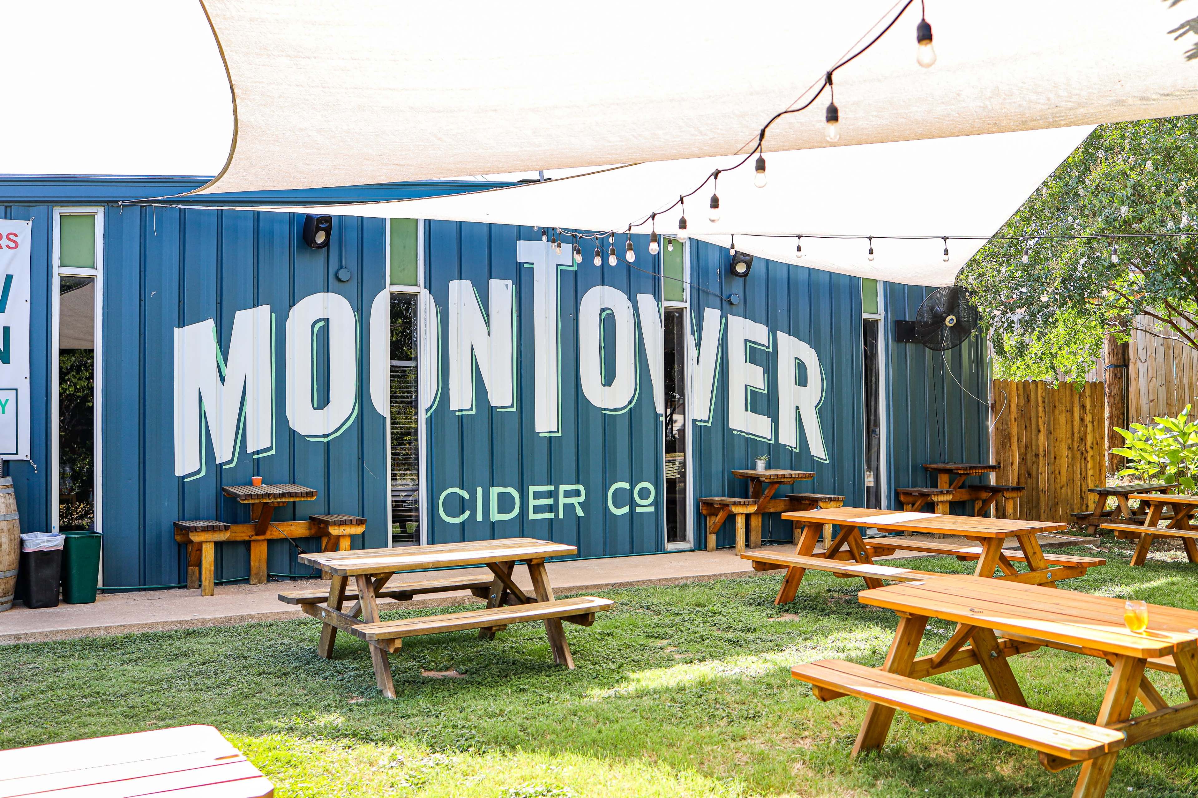 The exterior of a cider company, Moon Tower Cider Co., with picnic tables in a grassy area and a blue building featuring large, white lettering.
