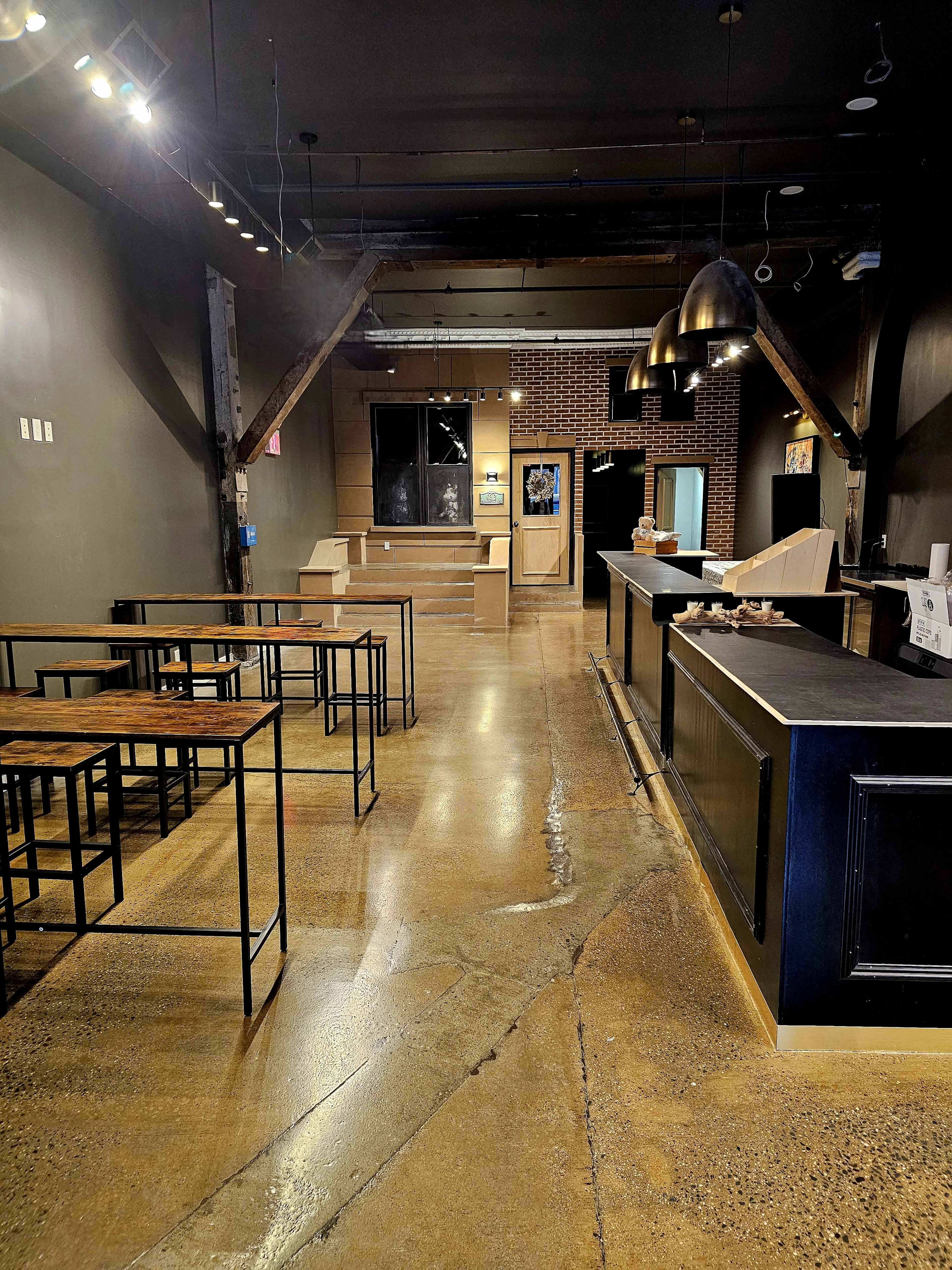The image shows a modern, empty cafe interior with a polished concrete floor, a sleek counter, and several tall tables arranged for seating.