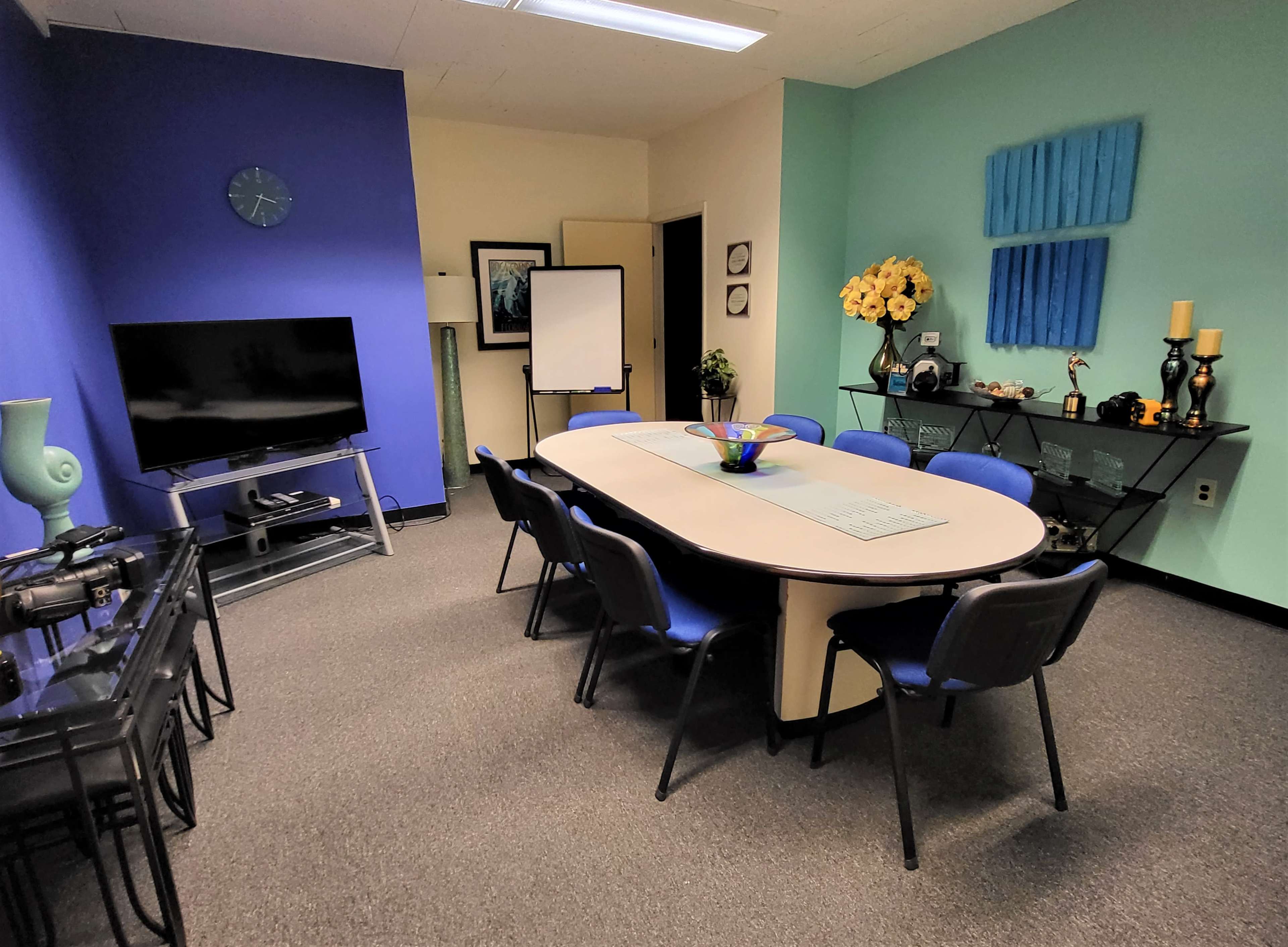 The image shows a meeting room with a large oval table surrounded by blue chairs, a television on a stand, and decorative artwork on the walls.