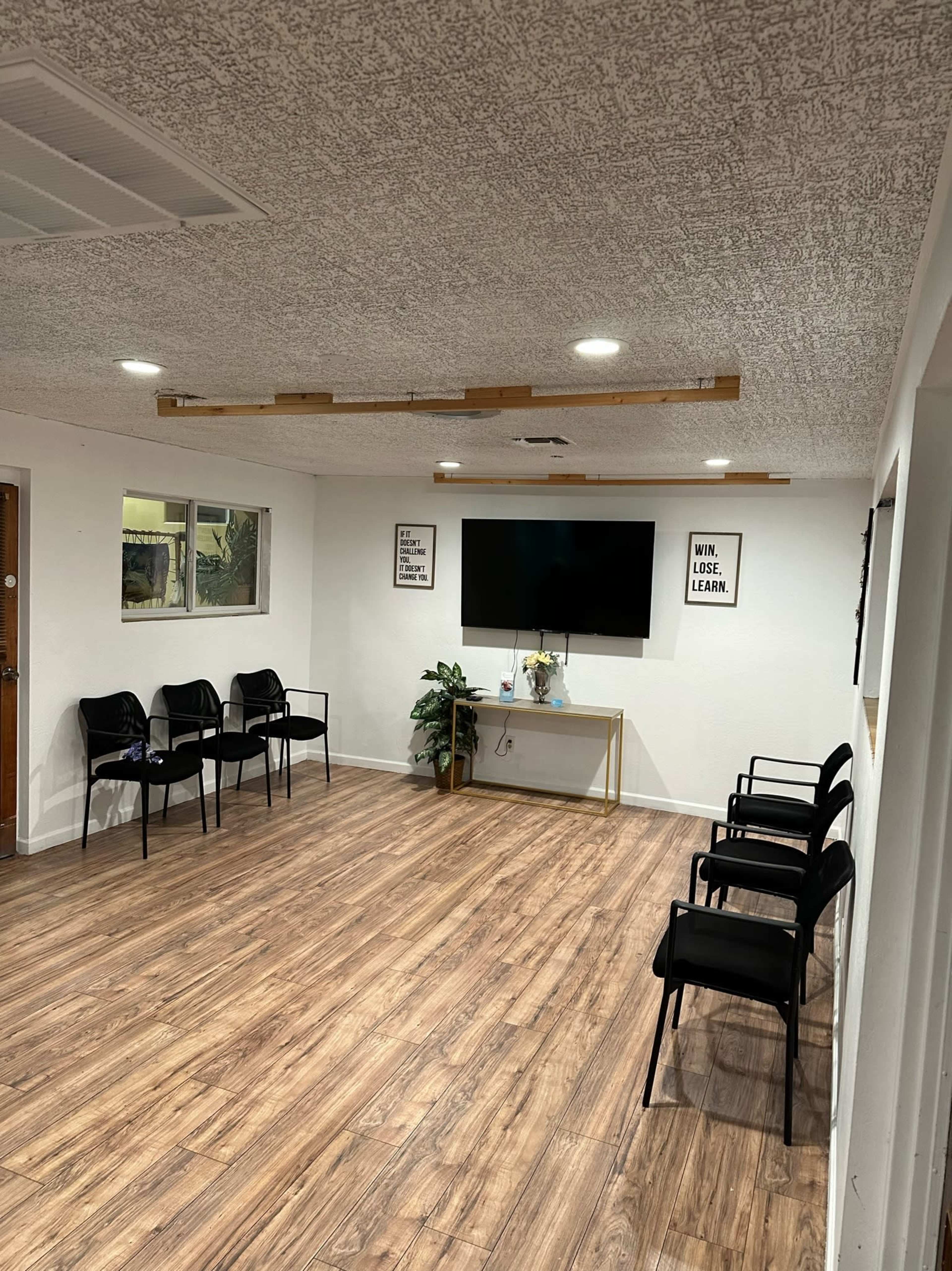 A spacious room with wooden flooring, featuring several black chairs arranged against the walls and a wall-mounted television opposite a small table with a plant.