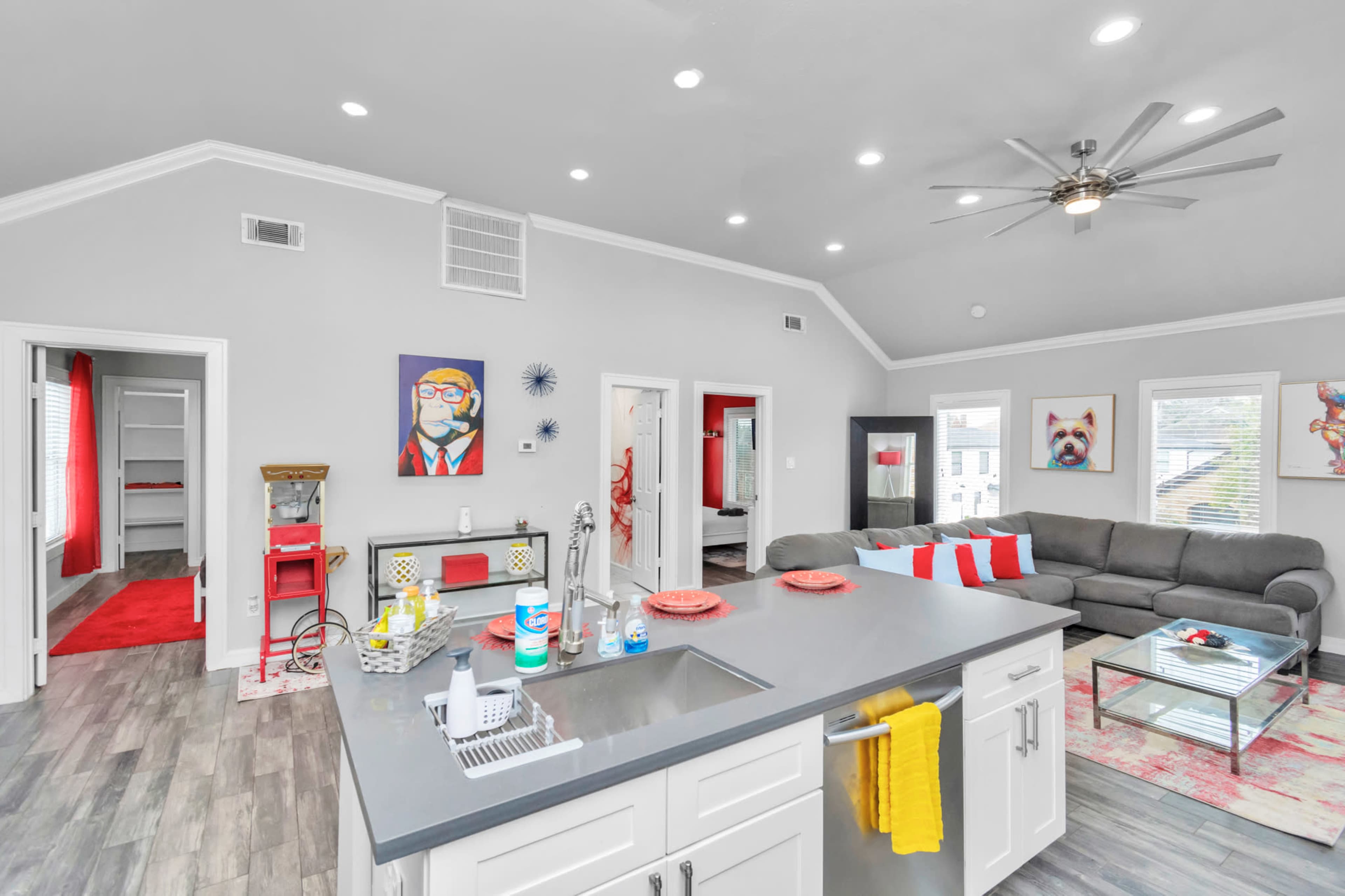 A modern open-concept living space featuring a kitchen island, a grey sectional sofa, colorful wall art, and bright accents in the decor.