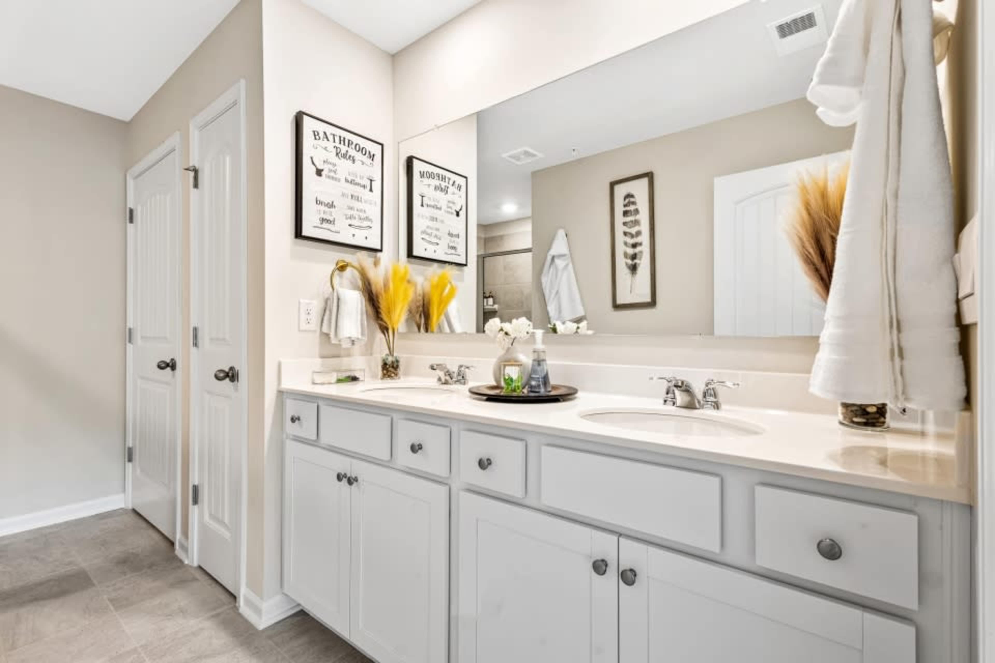 The image shows a well-lit bathroom featuring double sinks, a large mirror, and decorative wall art.