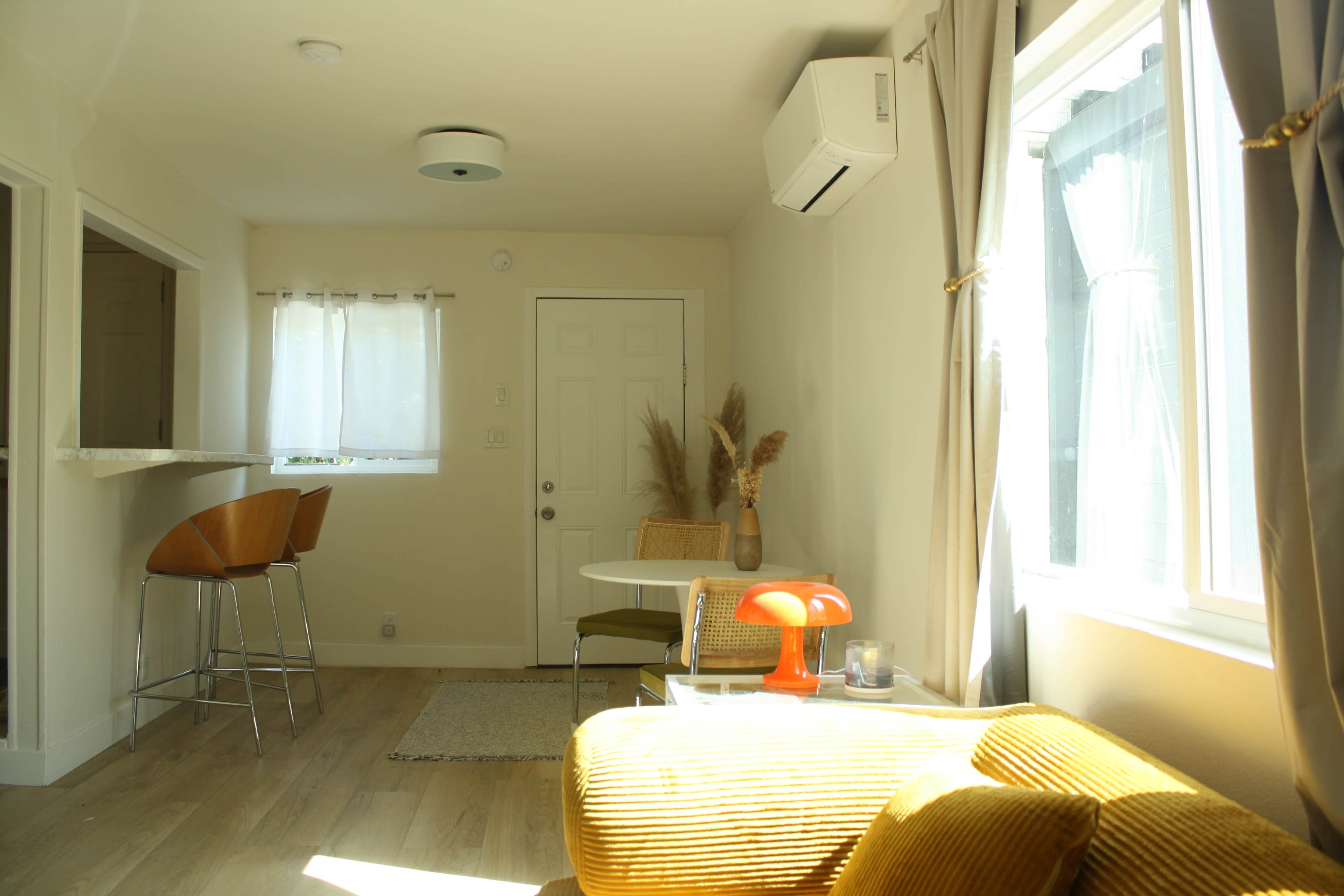 The image shows a bright, modern living area with a small kitchen bar, a dining table, a yellow couch, and large windows allowing natural light to enter.