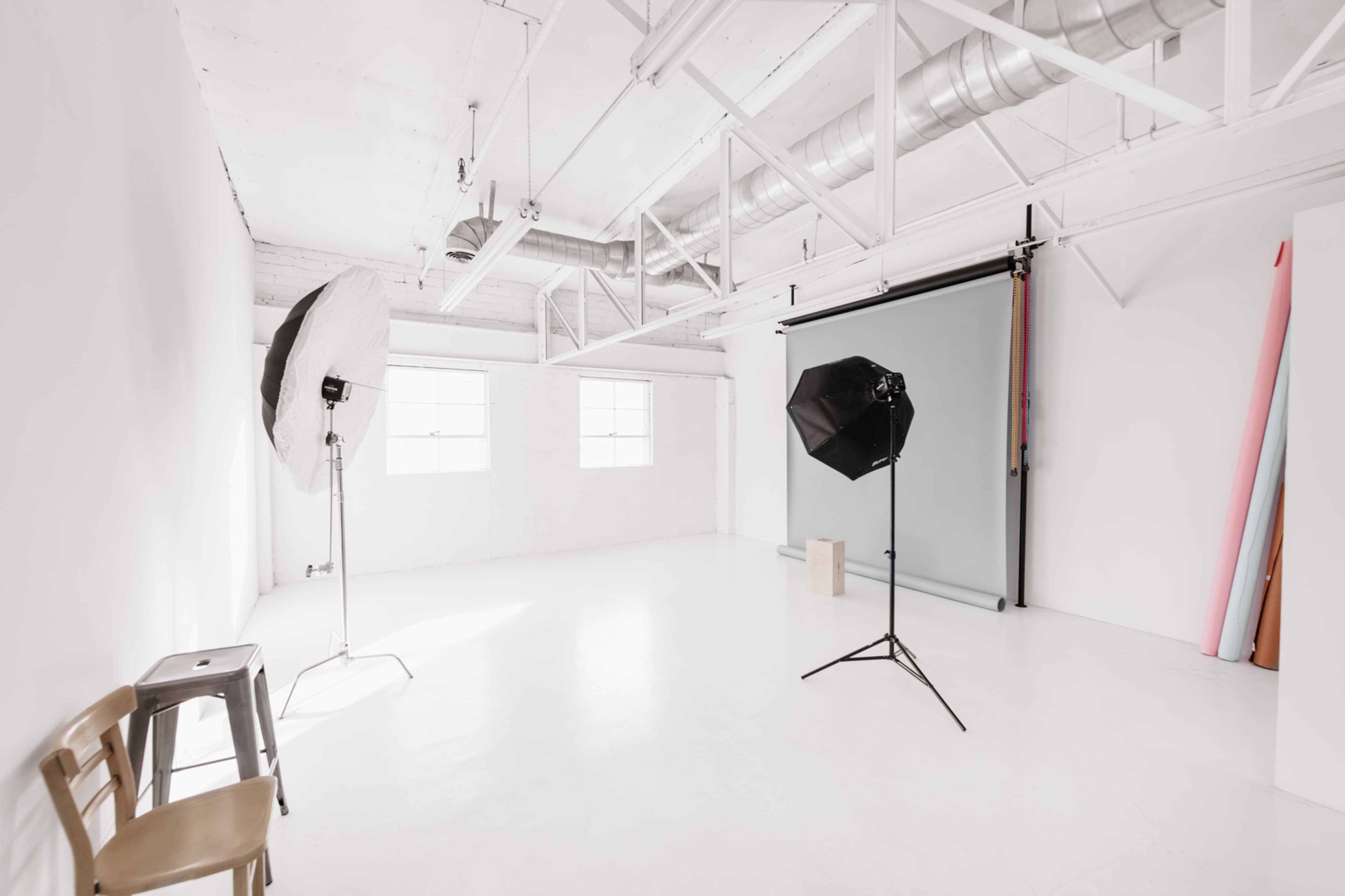 A spacious, well-lit photography studio with two light setup stands, a backdrop, and a few chairs.