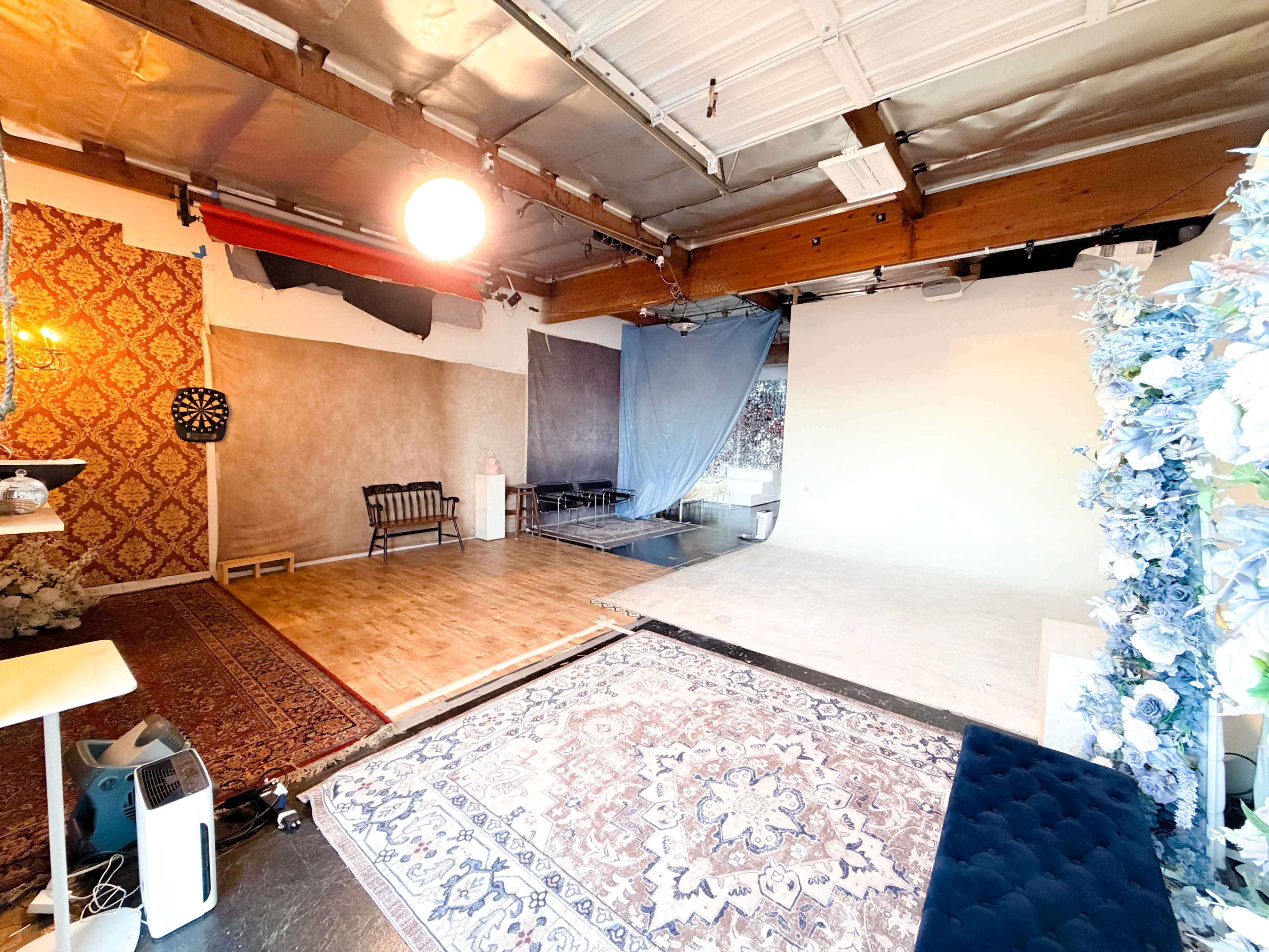 The image shows a spacious studio interior with wooden beams, various textured backdrops, and decorative rugs on the floor.