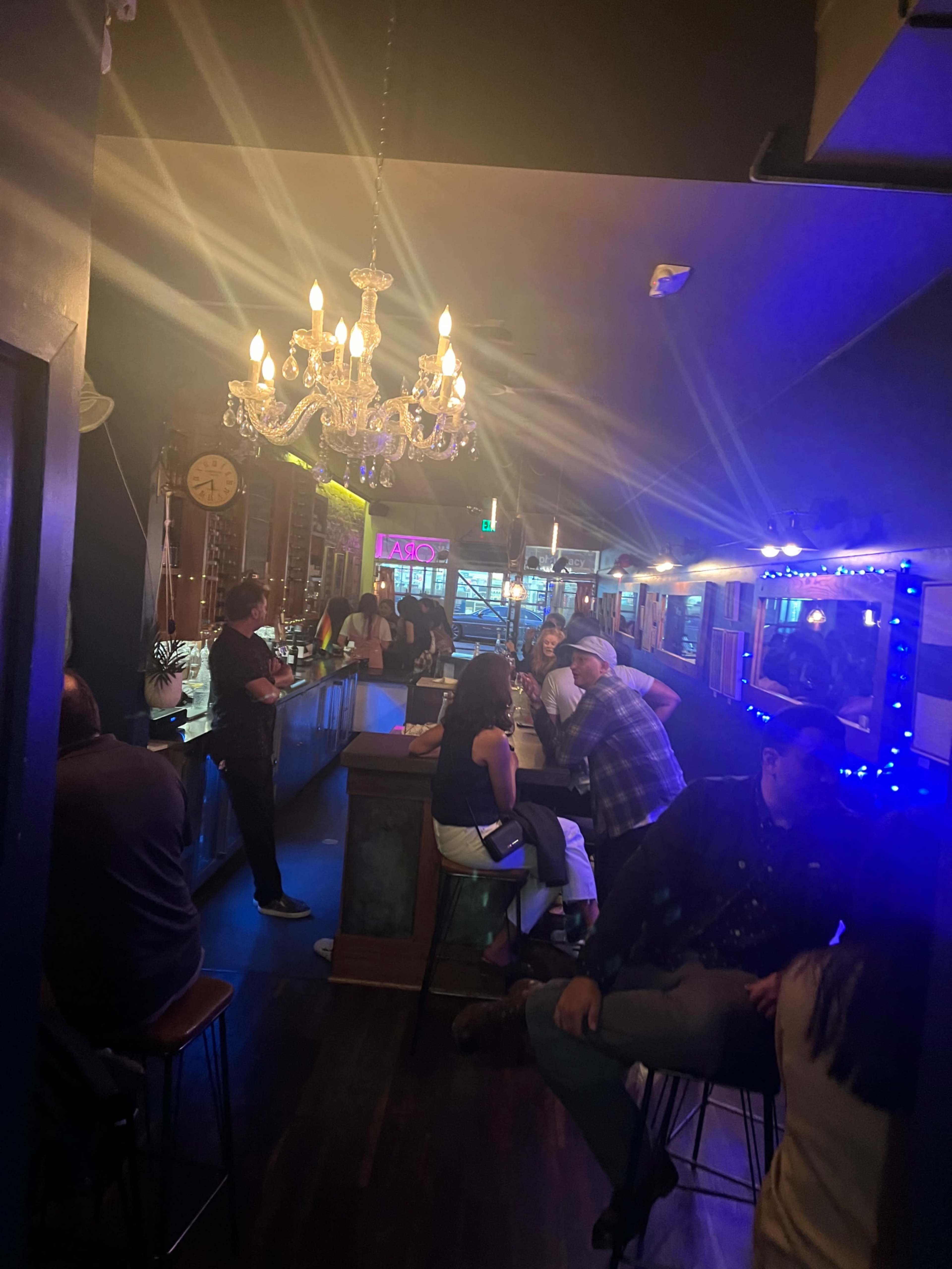 A dimly lit bar features patrons seated at the counter and various tables, with a chandelier hanging above and colorful lights illuminating the space.