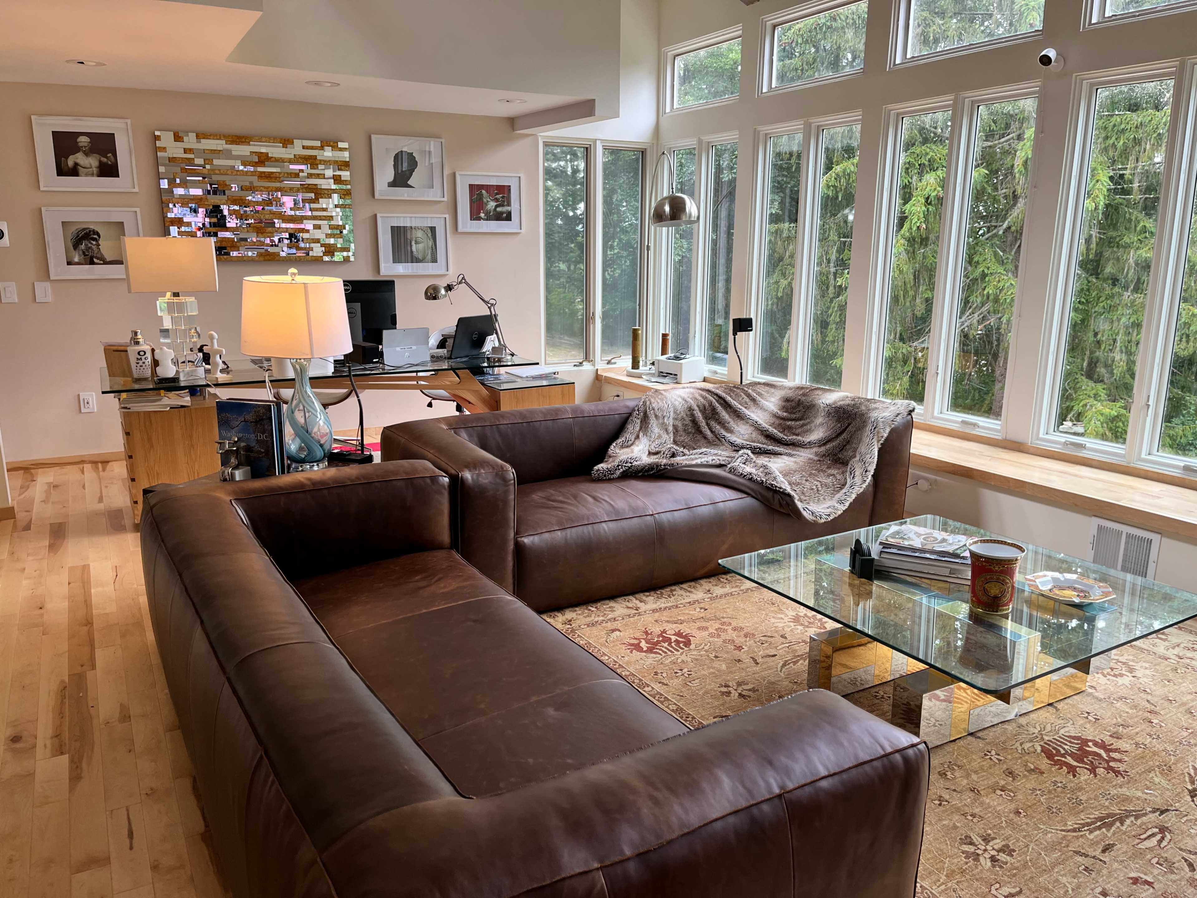 A cozy living room with two brown leather sofas, a glass coffee table, and large windows overlooking greenery, along with a desk area and decorative art on the walls.