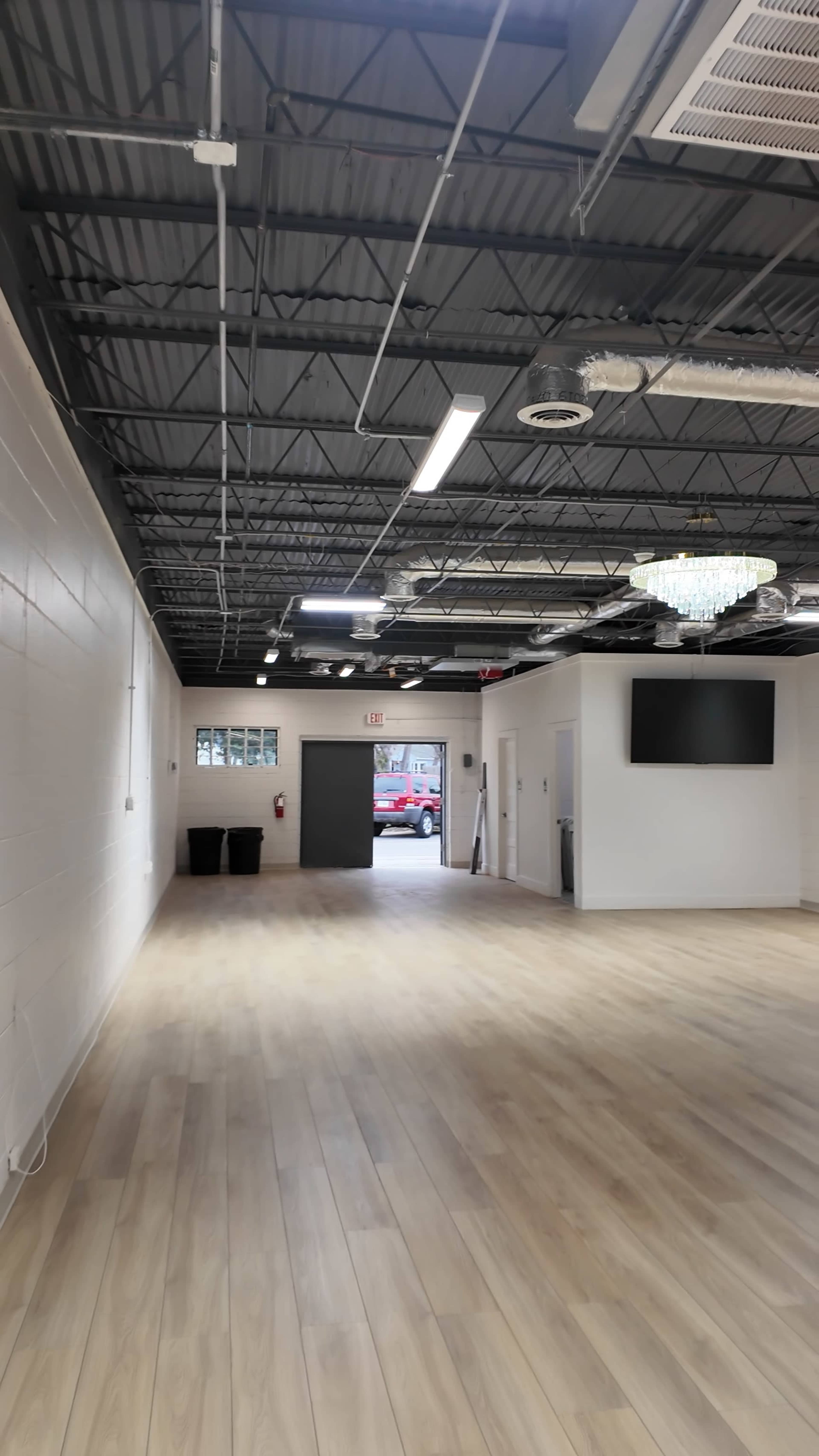 The image shows a spacious, well-lit interior of a building with a wooden floor, white walls, and a chandelier hanging from the ceiling, leading to an open doorway.
