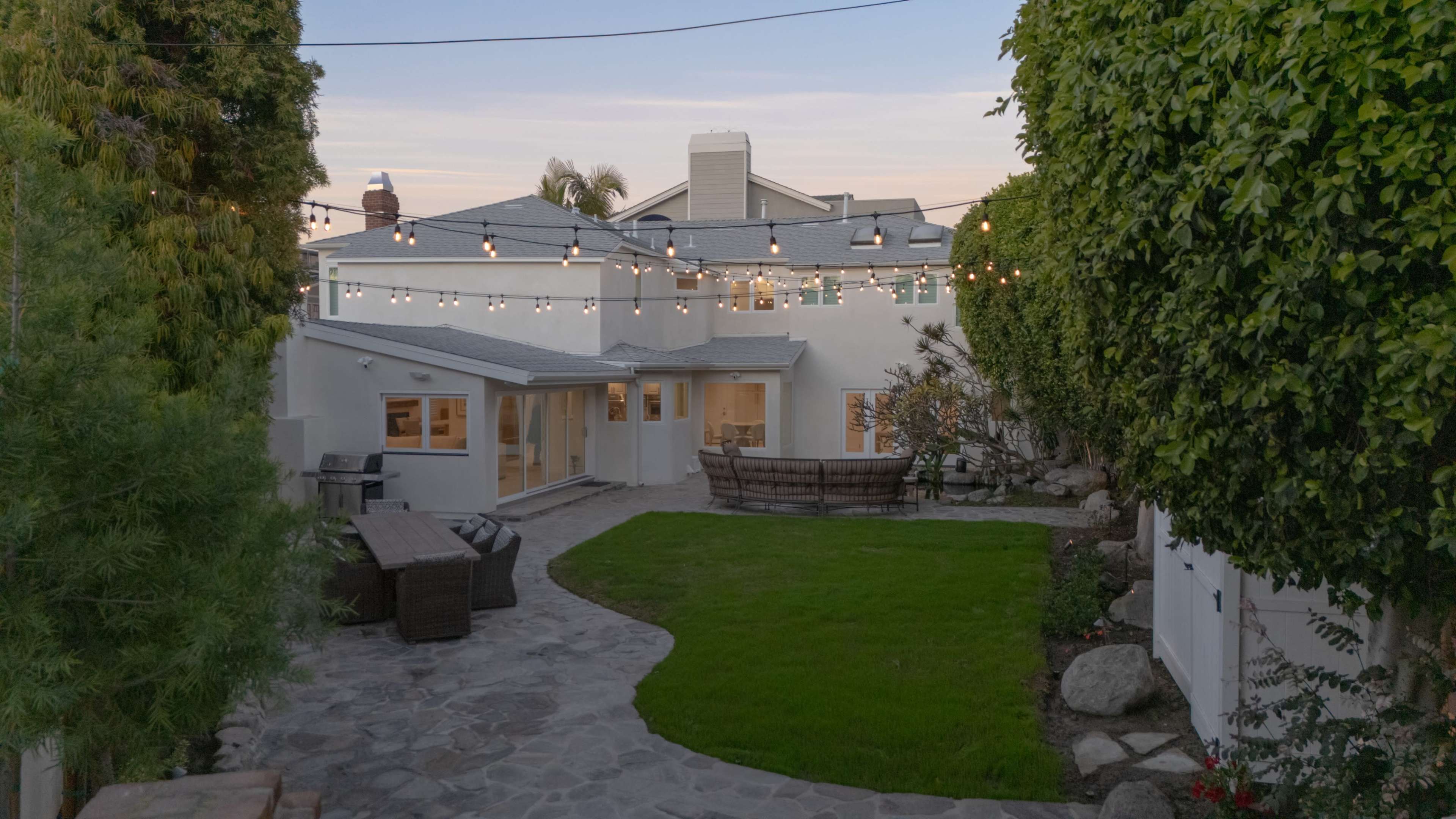 The image shows a well-maintained backyard featuring a stone pathway, a green lawn, and string lights overhead illuminating a seating area.