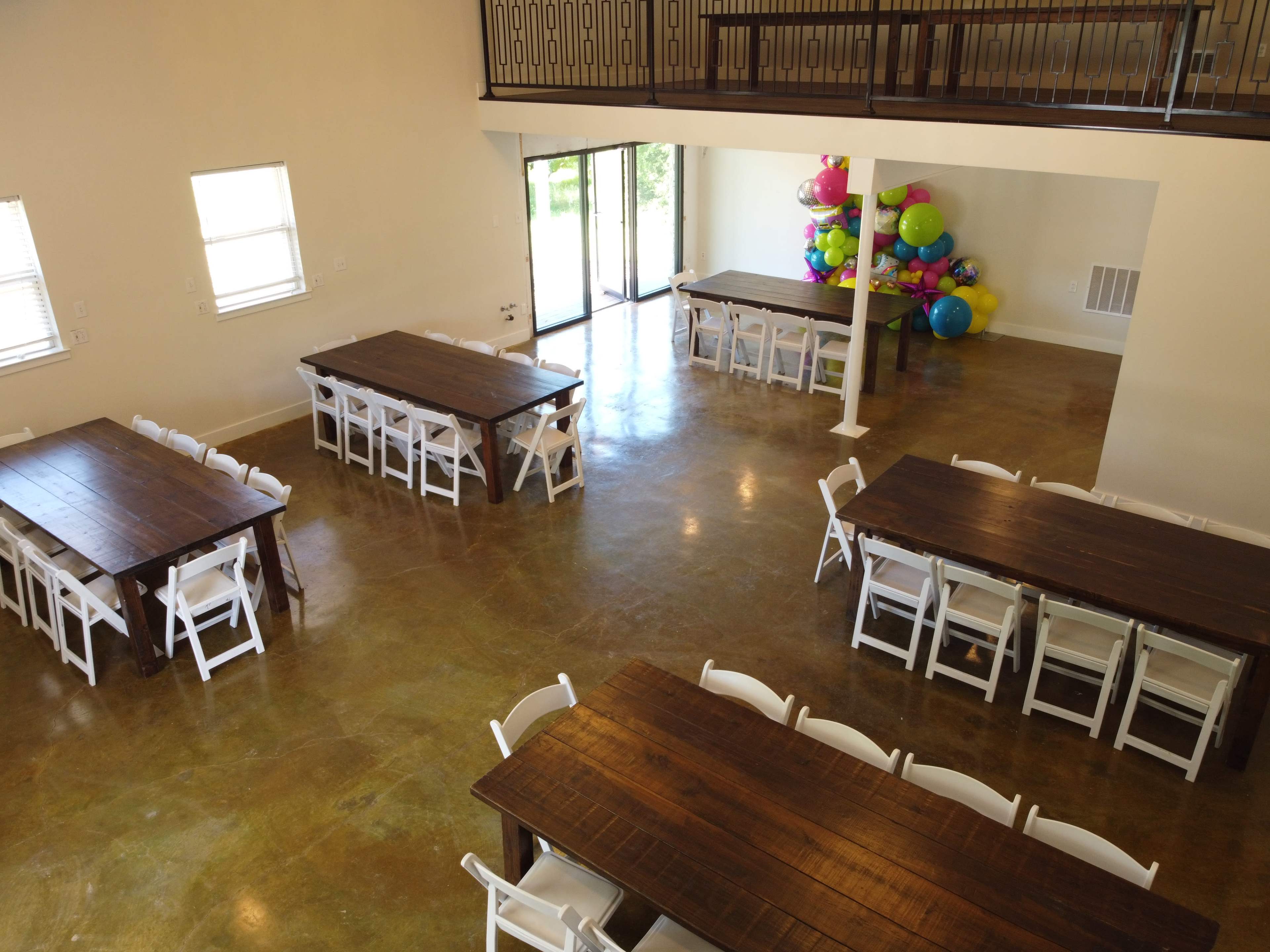 A spacious event room with multiple wooden tables arranged in rows and a cluster of colorful balloons in the corner.