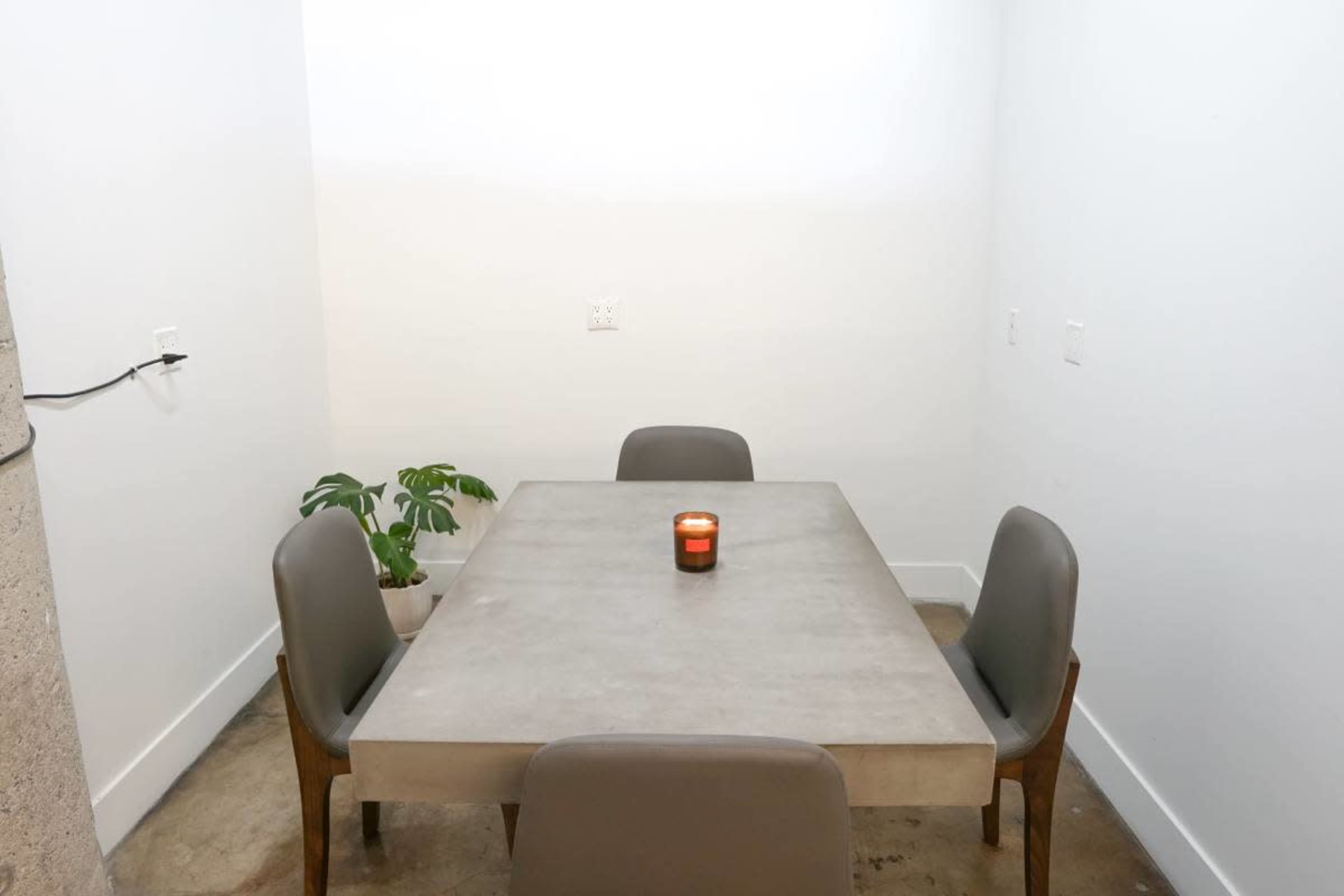 A small, minimalistic dining area features a concrete table with four chairs, a candle in the center, and a potted plant in the corner.