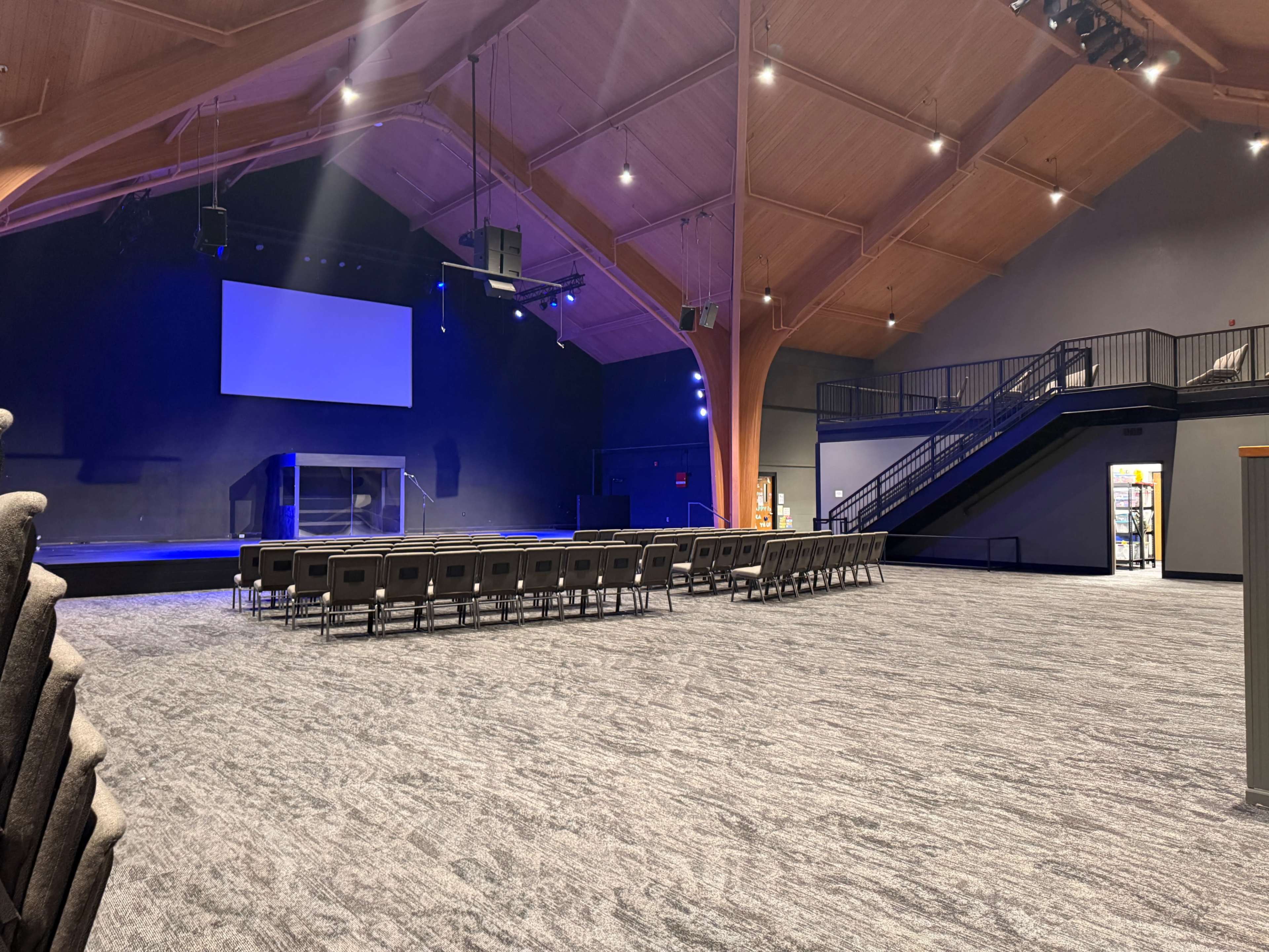 The image shows a spacious auditorium with a stage, rows of chairs arranged for an audience, and wooden beams supporting the ceiling.
