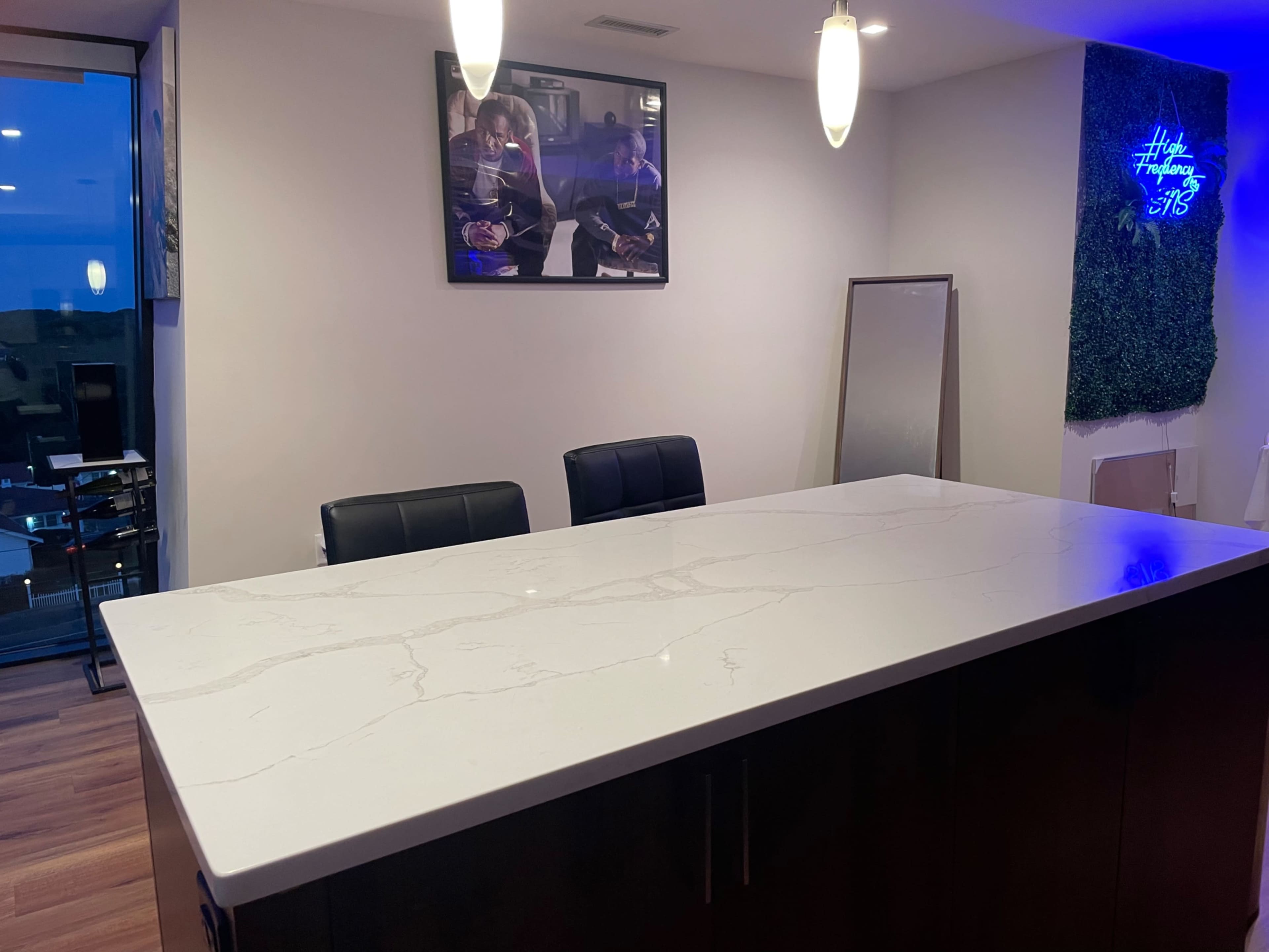 A modern kitchen features a large, white marble countertop with two black chairs set against a wall displaying a framed photo and a decorative green panel.