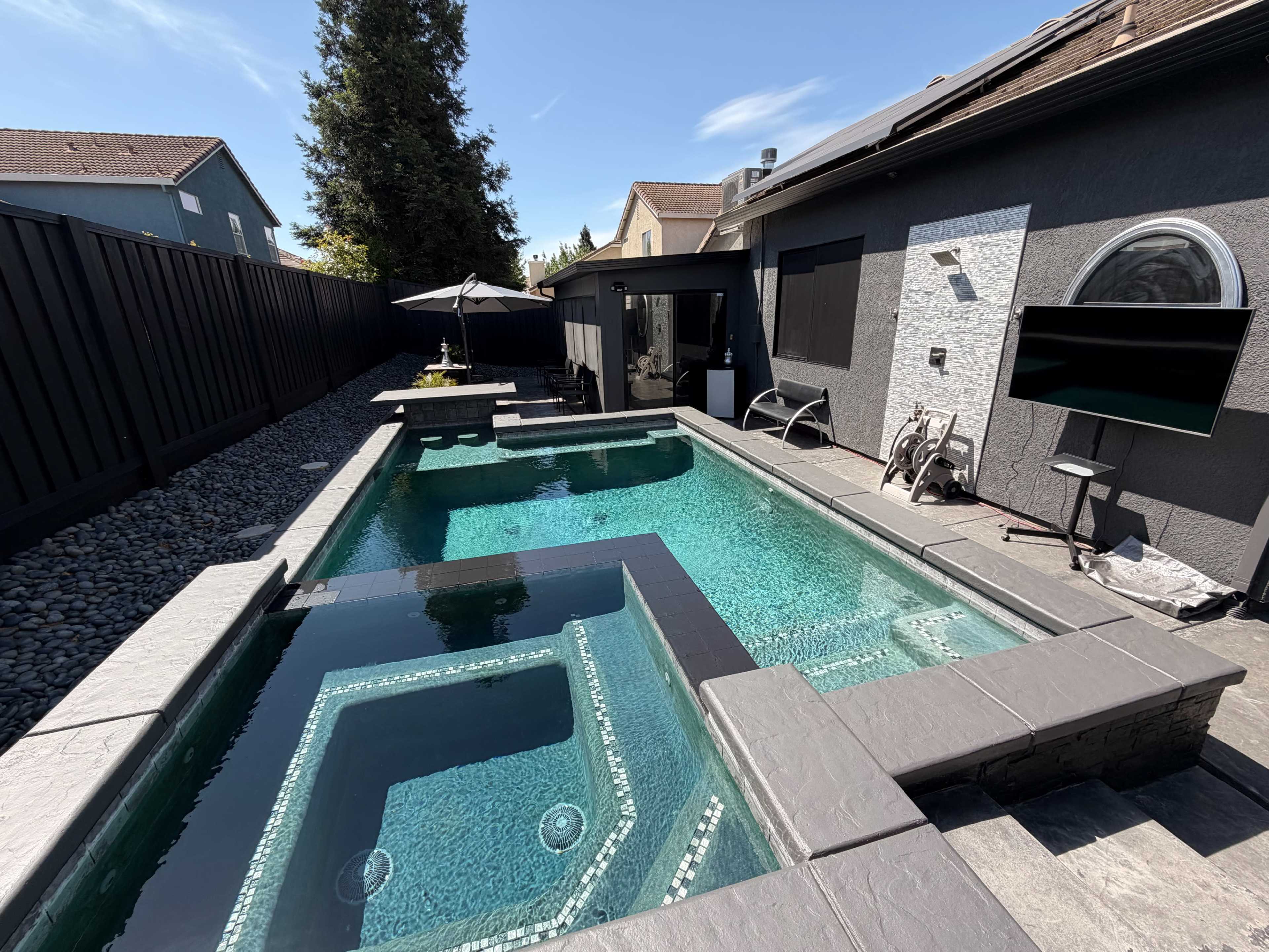 The image shows a modern backyard with a rectangular swimming pool surrounded by a stone patio, featuring a hot tub section and a black fence in the background.