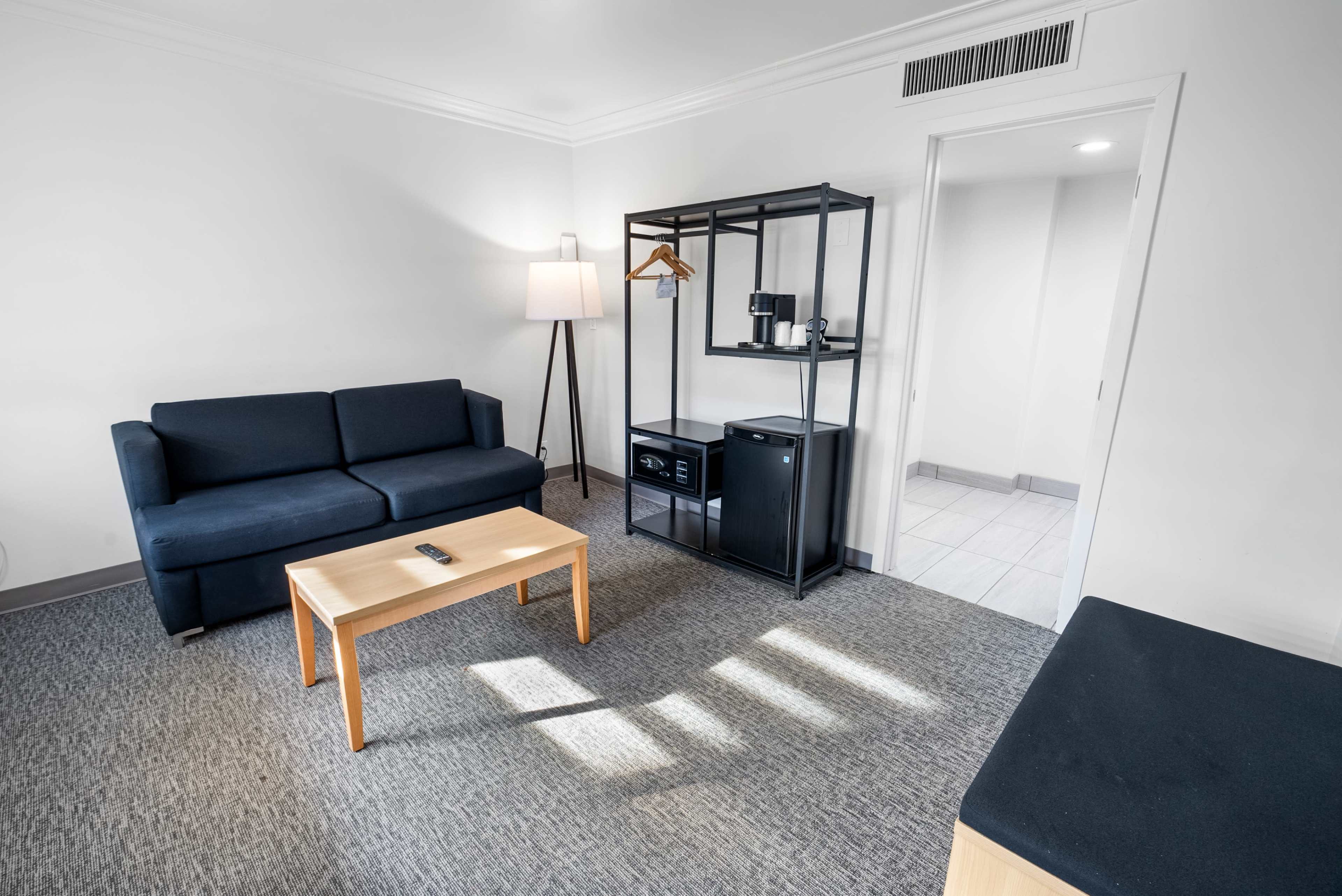 A minimalist hotel room with a black sofa, a wooden coffee table, and a shelving unit containing a coffee maker and a mini-fridge.