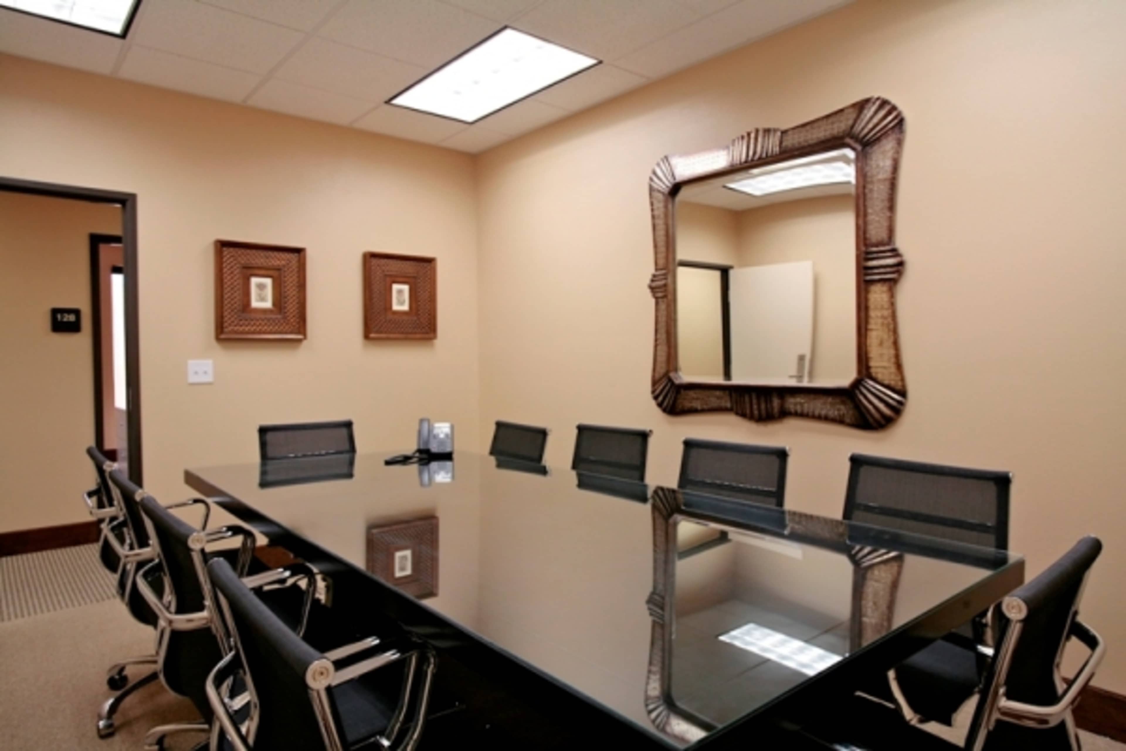 A conference room features a long glass table surrounded by black chairs, with decorative frames on the walls and a mirrored wall reflecting the space.