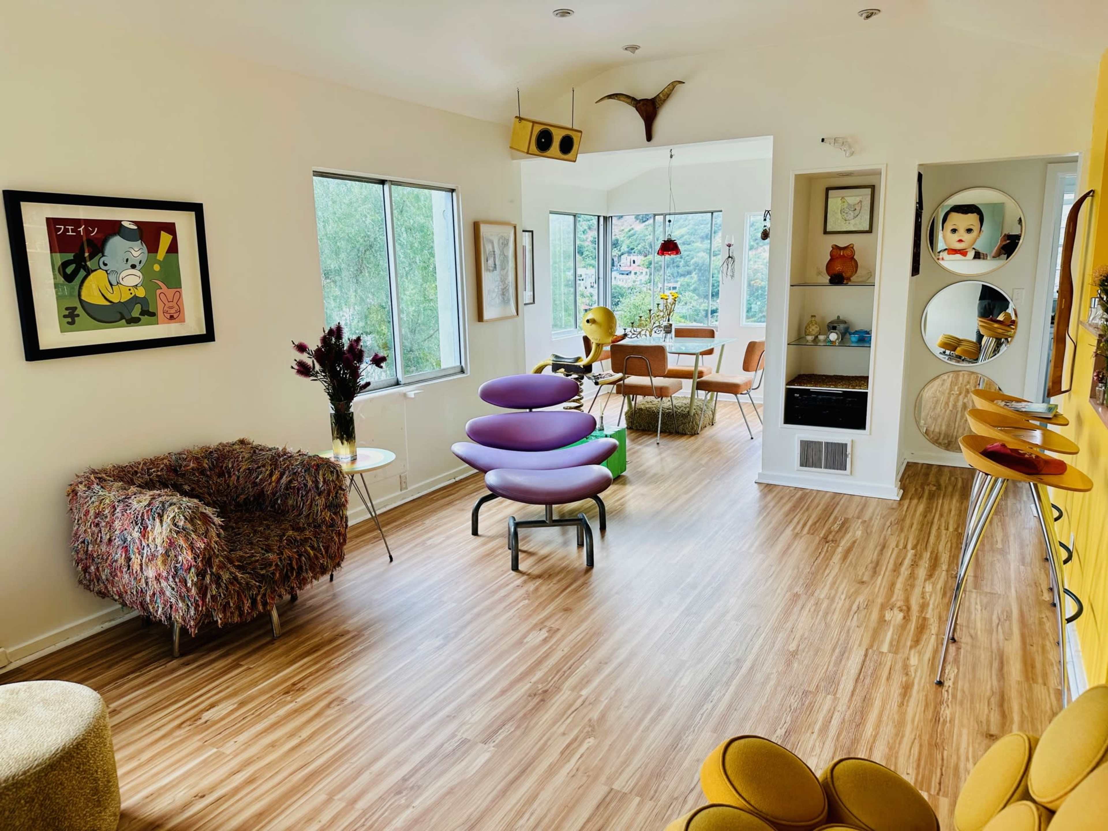 The image shows a modern living room with wooden floors, featuring colorful furniture, large windows, and a mix of decorative elements.
